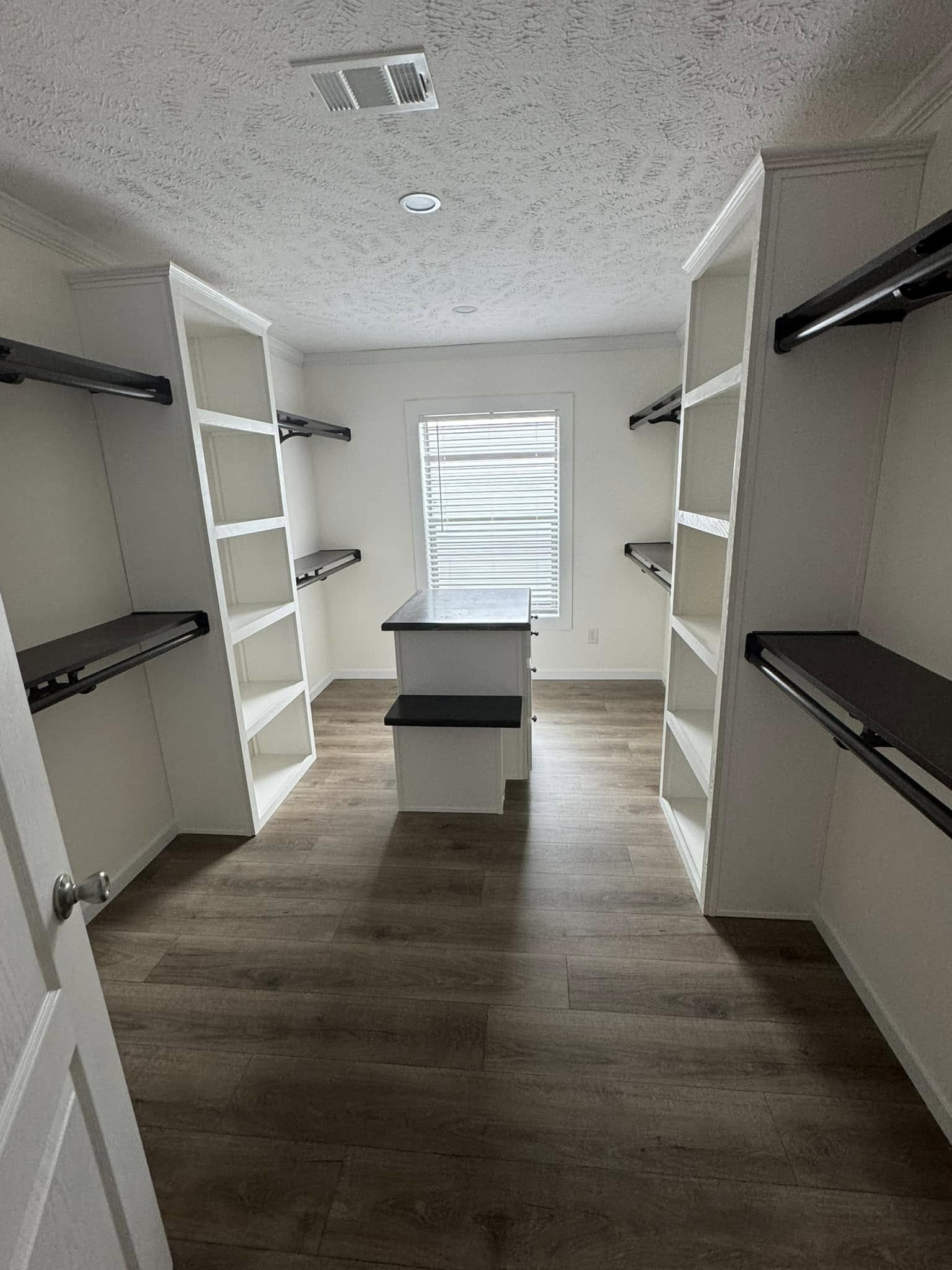 Spacious walk-in closet with white shelving, dark rods, and a central island beneath a window. Wood floor and bright lighting create a clean, organized feel.