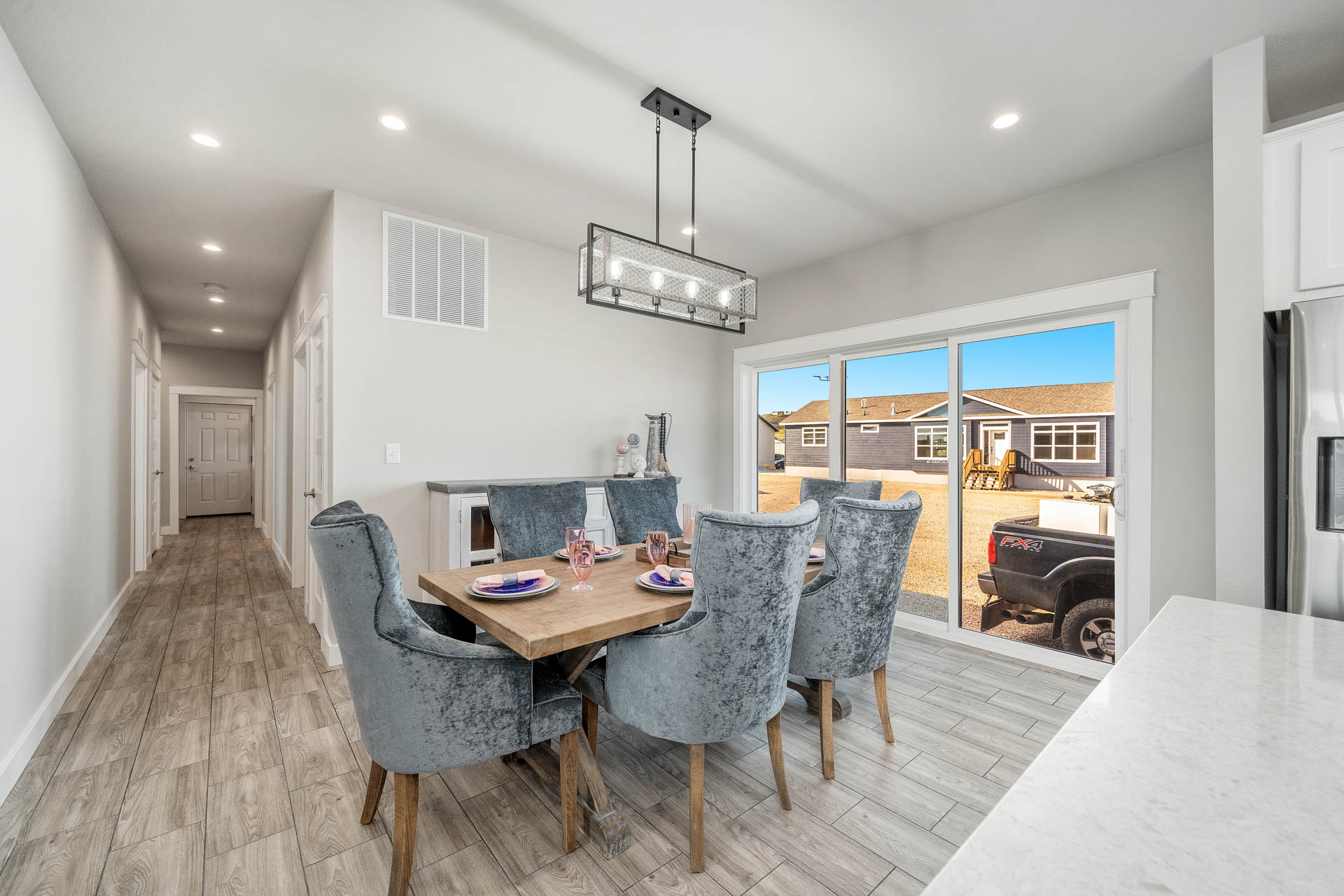 Modern dining area with a light wooden table and plush gray chairs set for six. Large windows overlook a sunny yard. Bright, minimalist decor.