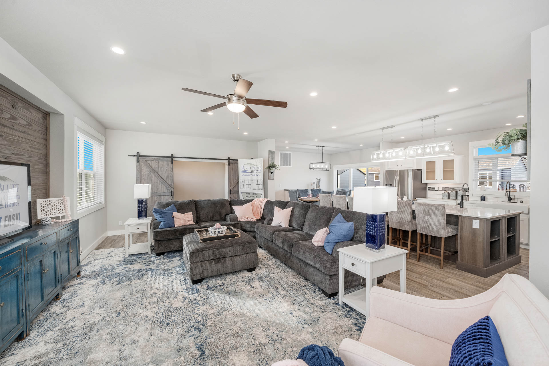 Spacious open-plan living room and kitchen with gray sofas, blue and pink cushions, patterned rug, wood accents, and modern pendant lighting. Cozy and inviting.
