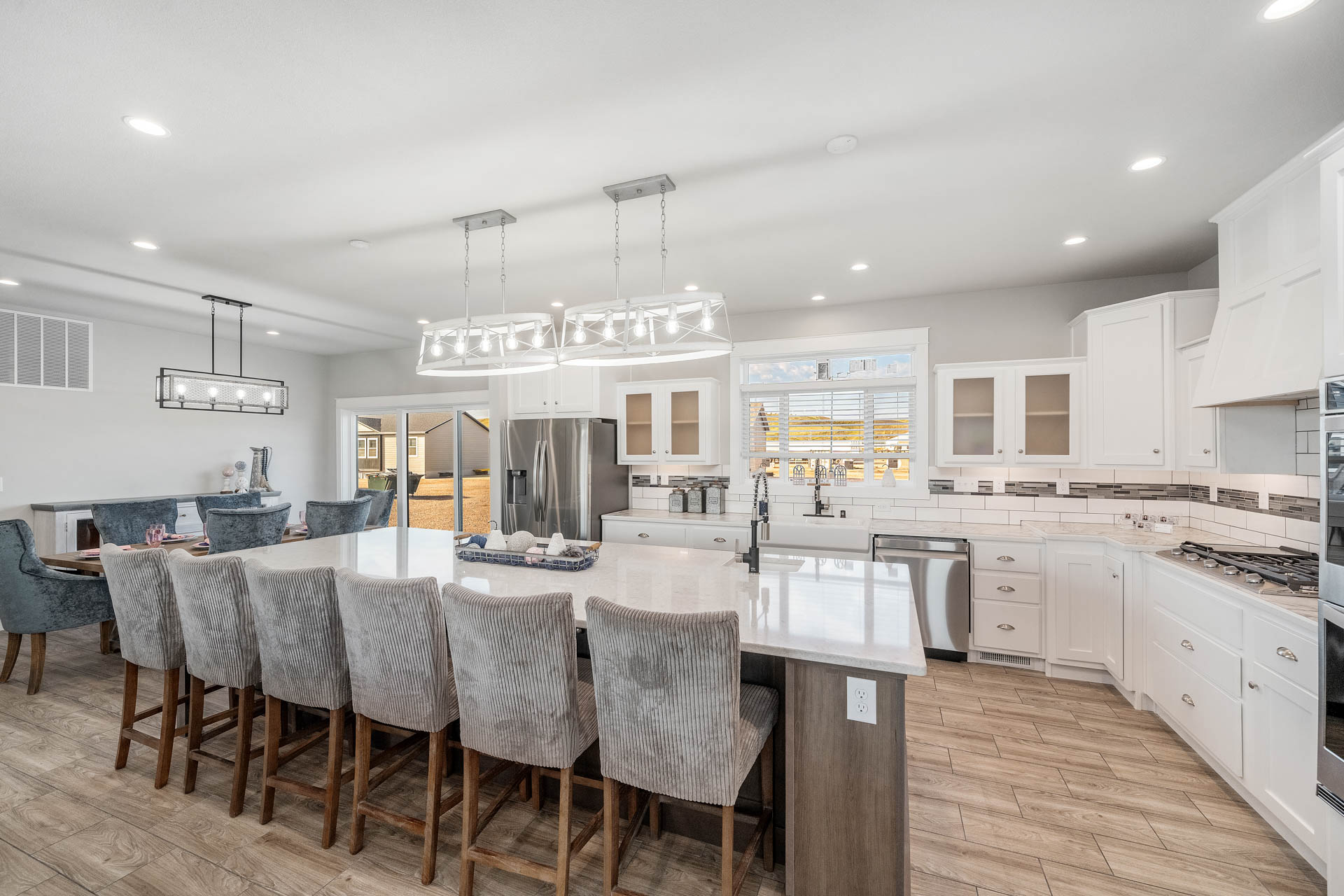 Spacious modern kitchen with white cabinets, stainless steel appliances, and a large island with six upholstered chairs. Bright lighting and wood floors create a welcoming ambiance.