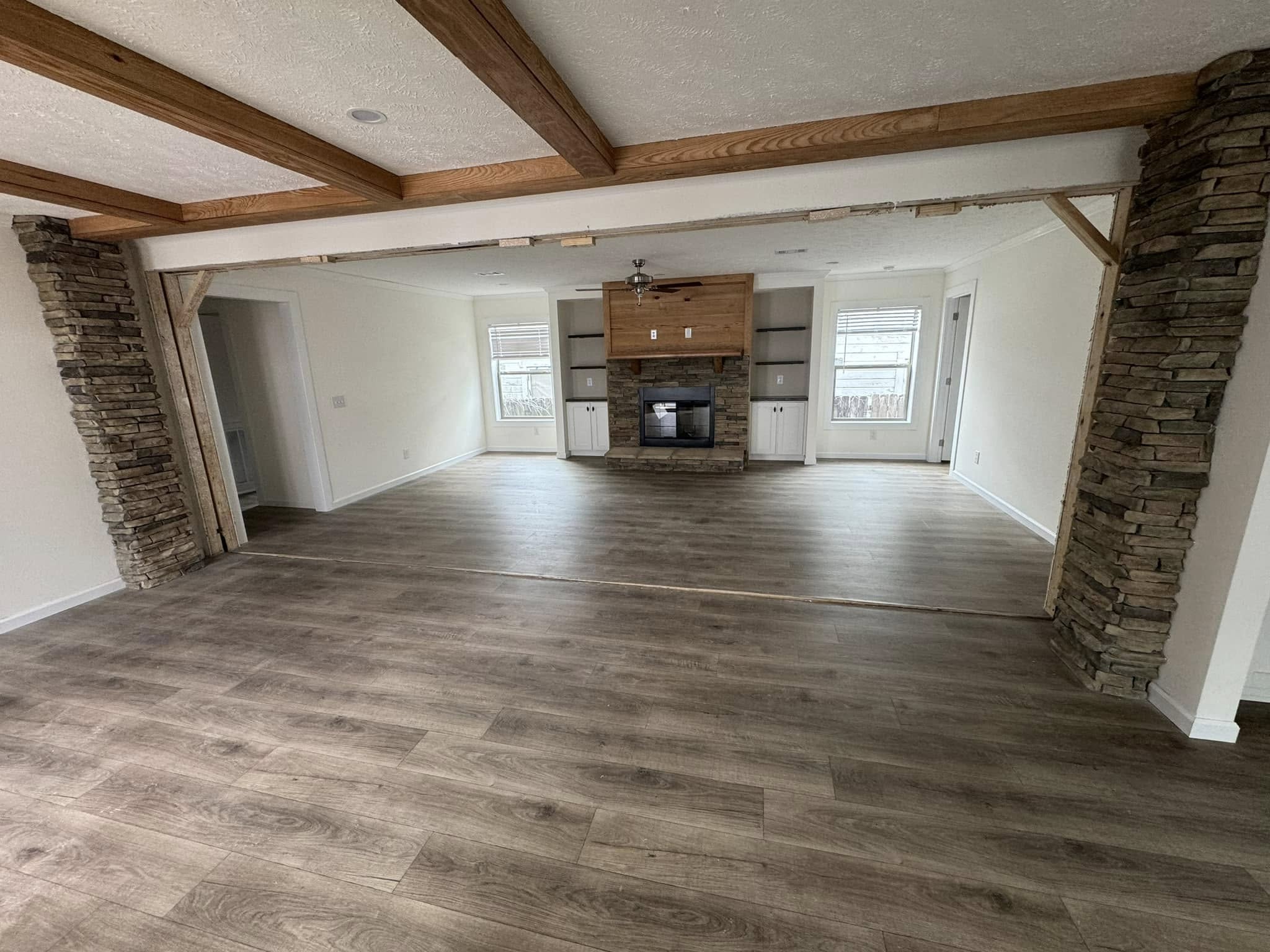 Spacious living room with light wood flooring, stone fireplace, and wooden ceiling beams. Large windows illuminate the inviting, rustic space.