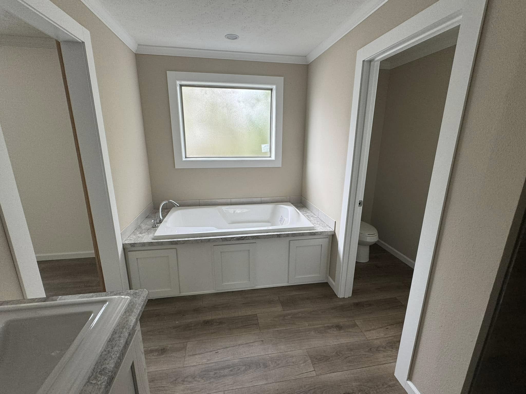 Modern bathroom with a white bathtub beneath a frosted window. Beige walls, wood-style flooring, and a visible small toilet room create a clean, serene atmosphere.