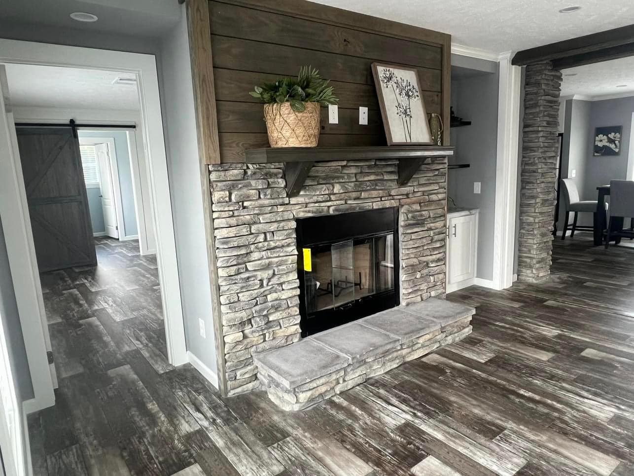 Rustic living room with a stone fireplace, wooden mantel, and wood-style flooring. Decor includes a plant and framed art. Calm and cozy ambiance.
