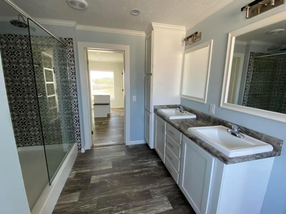 Bright bathroom with a double sink vanity, mirrored cabinets, and patterned tile shower. Wood-like flooring and soft blue-gray walls give a calm, modern feel.