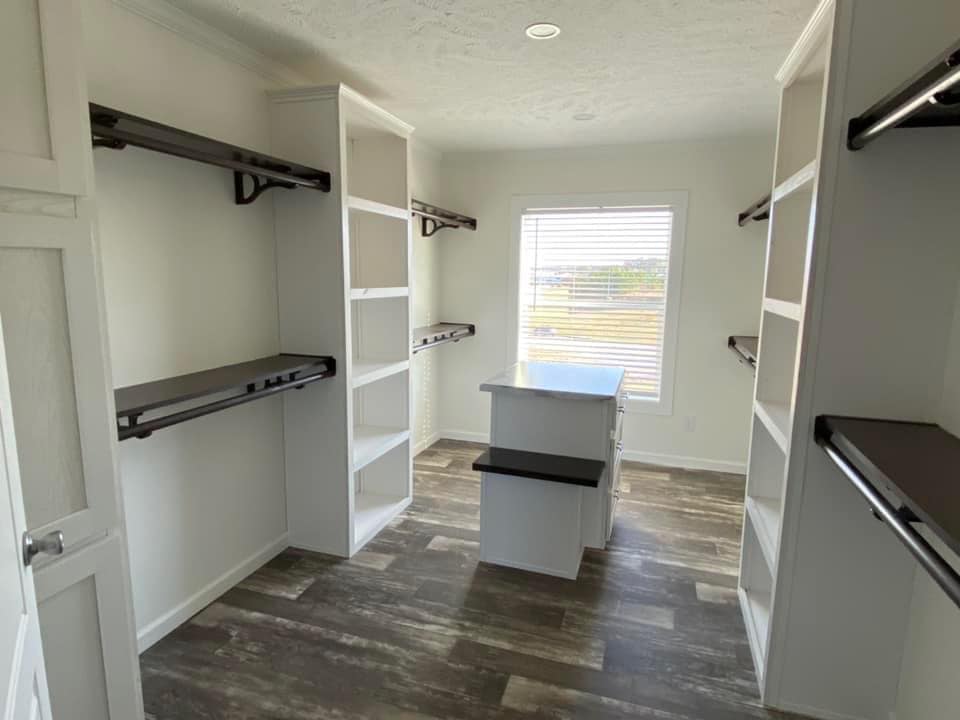 Spacious walk-in closet with white shelves and dark rods on both sides, central island, wood flooring, and a sunlit window creating a bright atmosphere.