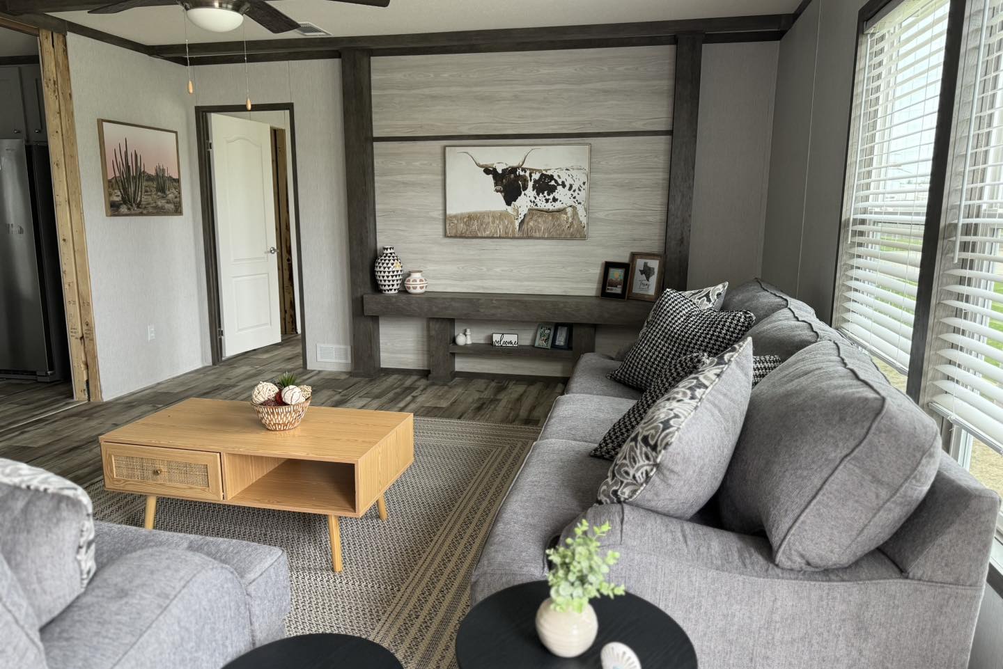 Cozy living room with gray sofas, patterned pillows, and a wooden coffee table. Art of a cow above a shelf; large windows bring in natural light.