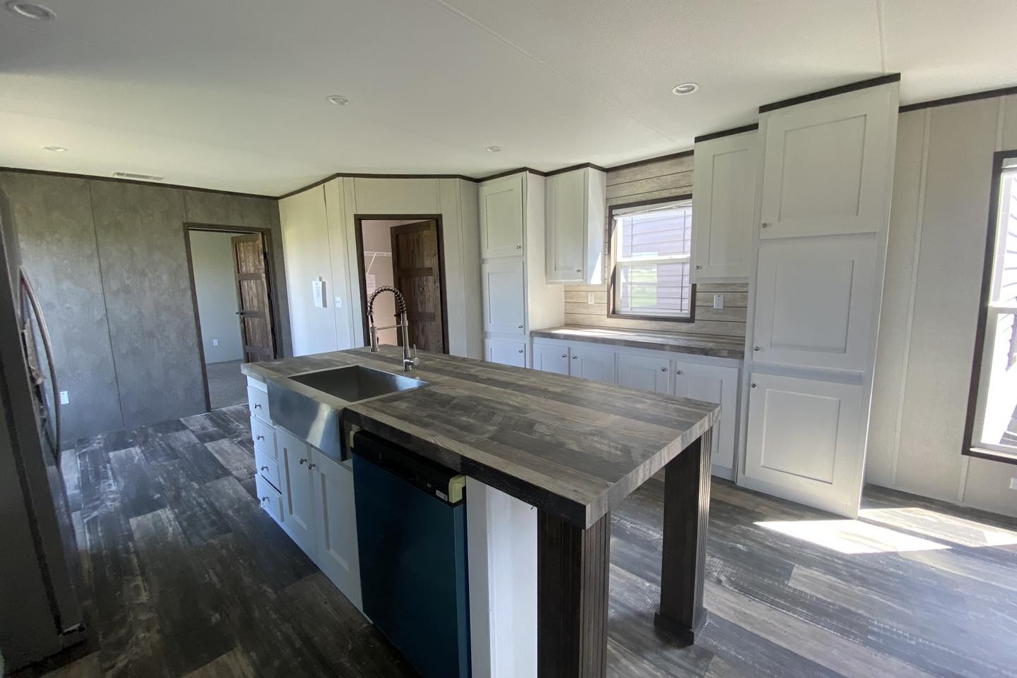 Modern kitchen with a large island featuring a sink and dishwasher. White cabinets, wood accents, and sunlight through windows create a bright, open atmosphere.
