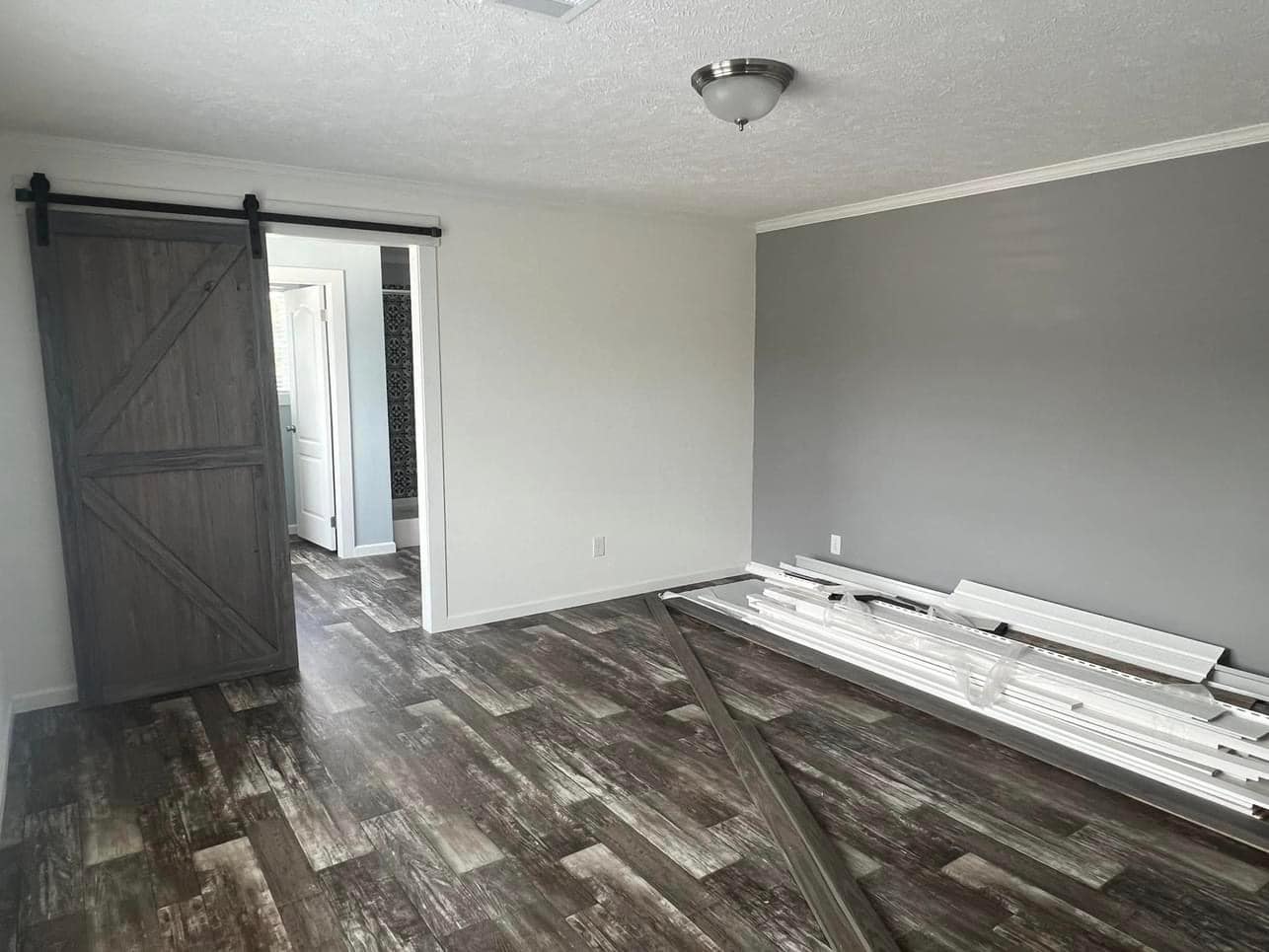 Empty room with gray barn door, dark wood floor, and two-tone gray walls. Stacked white trim boards and tool indicate ongoing renovation.