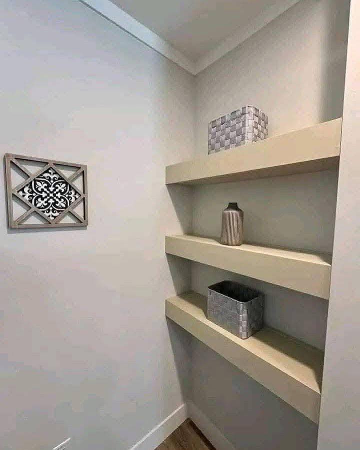 Simple corner with three beige floating shelves. Top shelf holds a woven box, middle has a tall vase, and bottom has a square basket. Wall art on left.