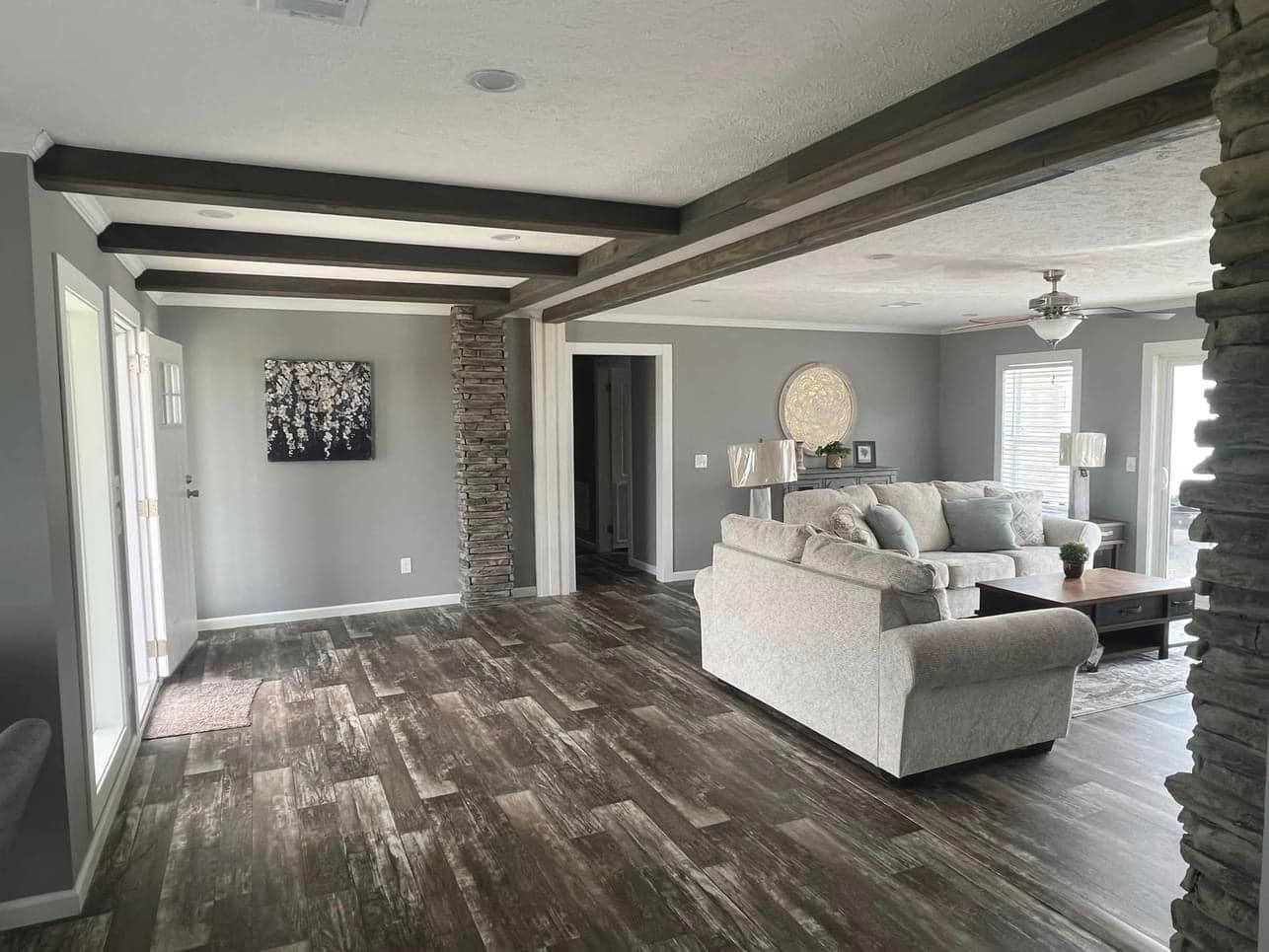 Spacious living room with gray walls, dark wooden beams, and stone pillars. Light gray sofas and a wooden coffee table create a cozy, modern feel.