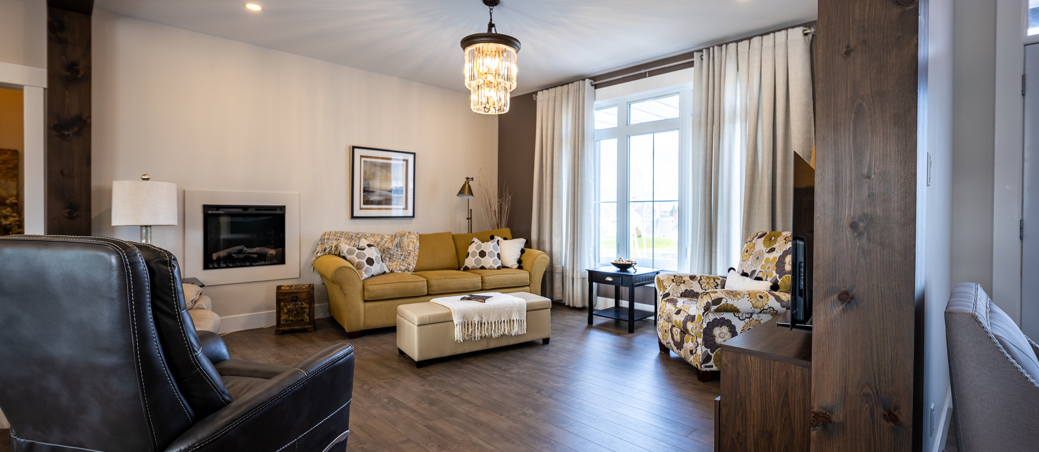 Cozy living room with a yellow sofa, floral armchair, and black leather recliner. A chandelier hangs from the ceiling, and large windows bring in natural light.