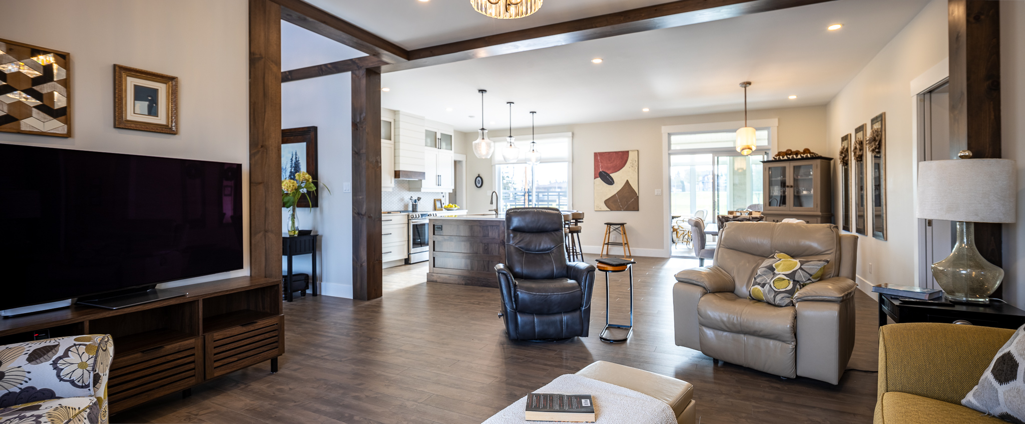 Modern open-plan living room and kitchen with wood flooring. Features leather recliners, a large TV, pendant lights, and abstract art. Cozy atmosphere.