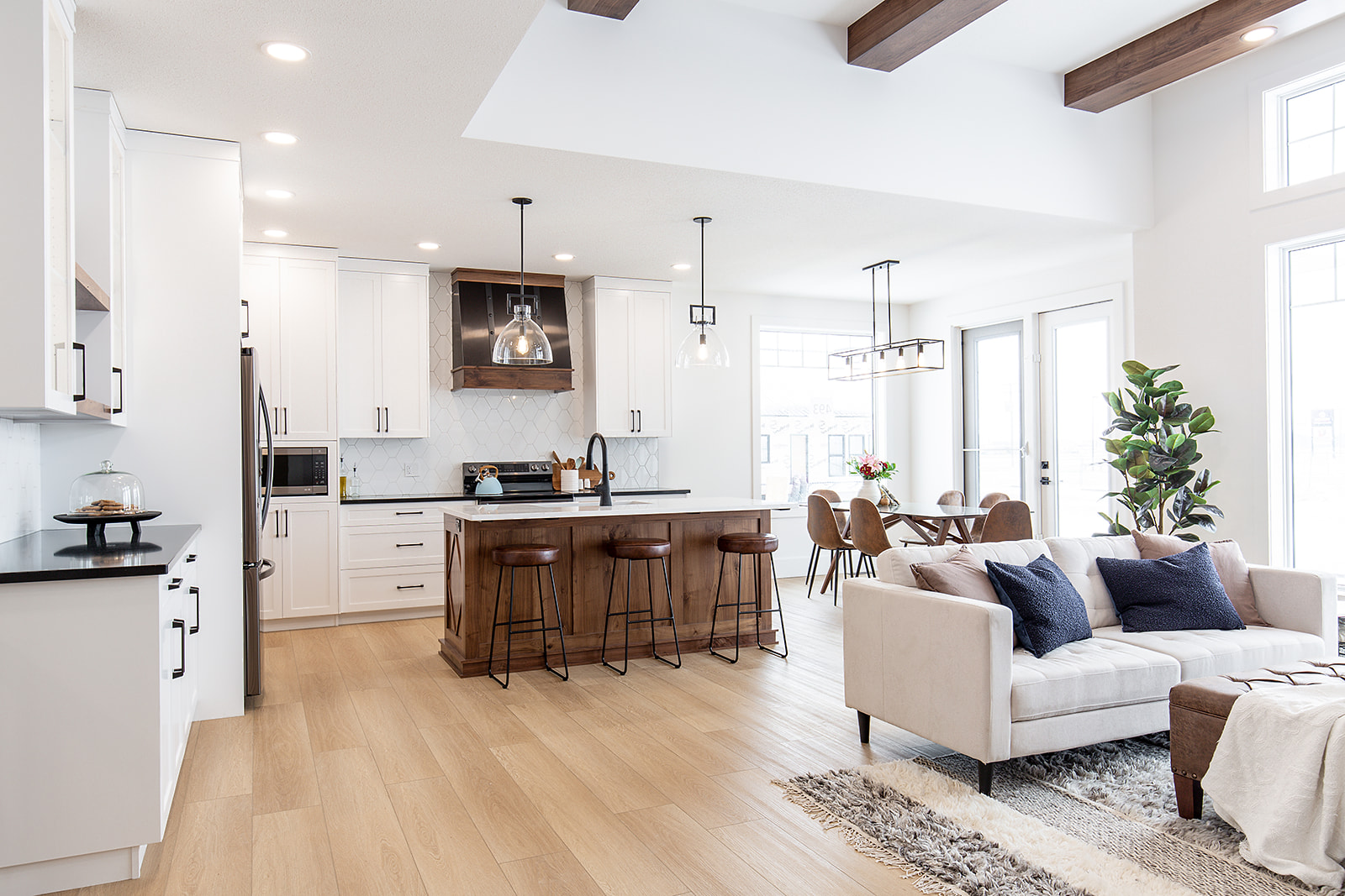 Modern open-plan kitchen and living area with wooden floors, white cabinets, dark wood island with stools, beige sofa, and large windows, creating a bright, inviting space.