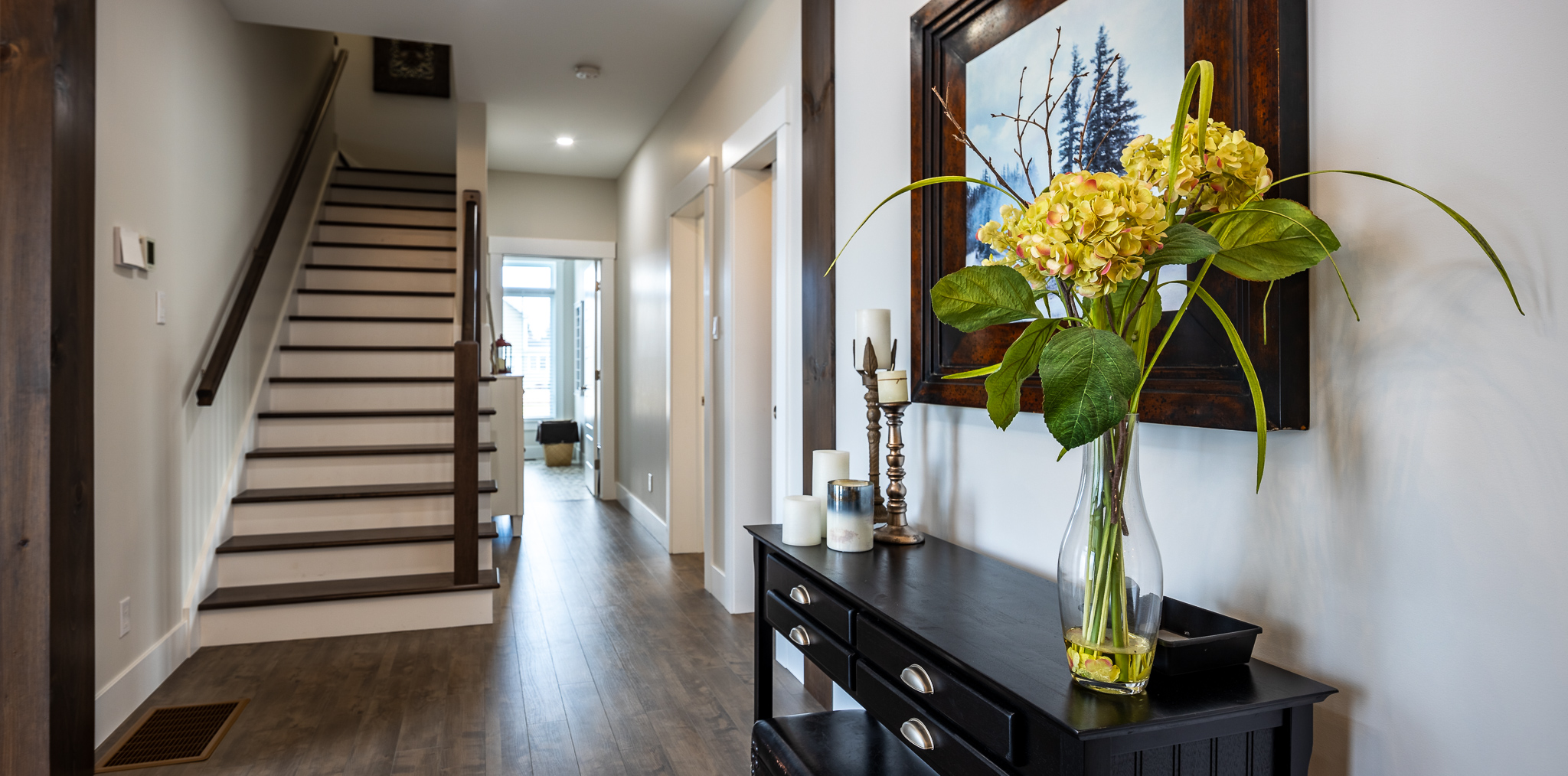 Spacious hallway with wooden floors, a staircase, and a black table adorned with a vase of yellow flowers. A landscape painting hangs above, creating a warm, welcoming ambiance.