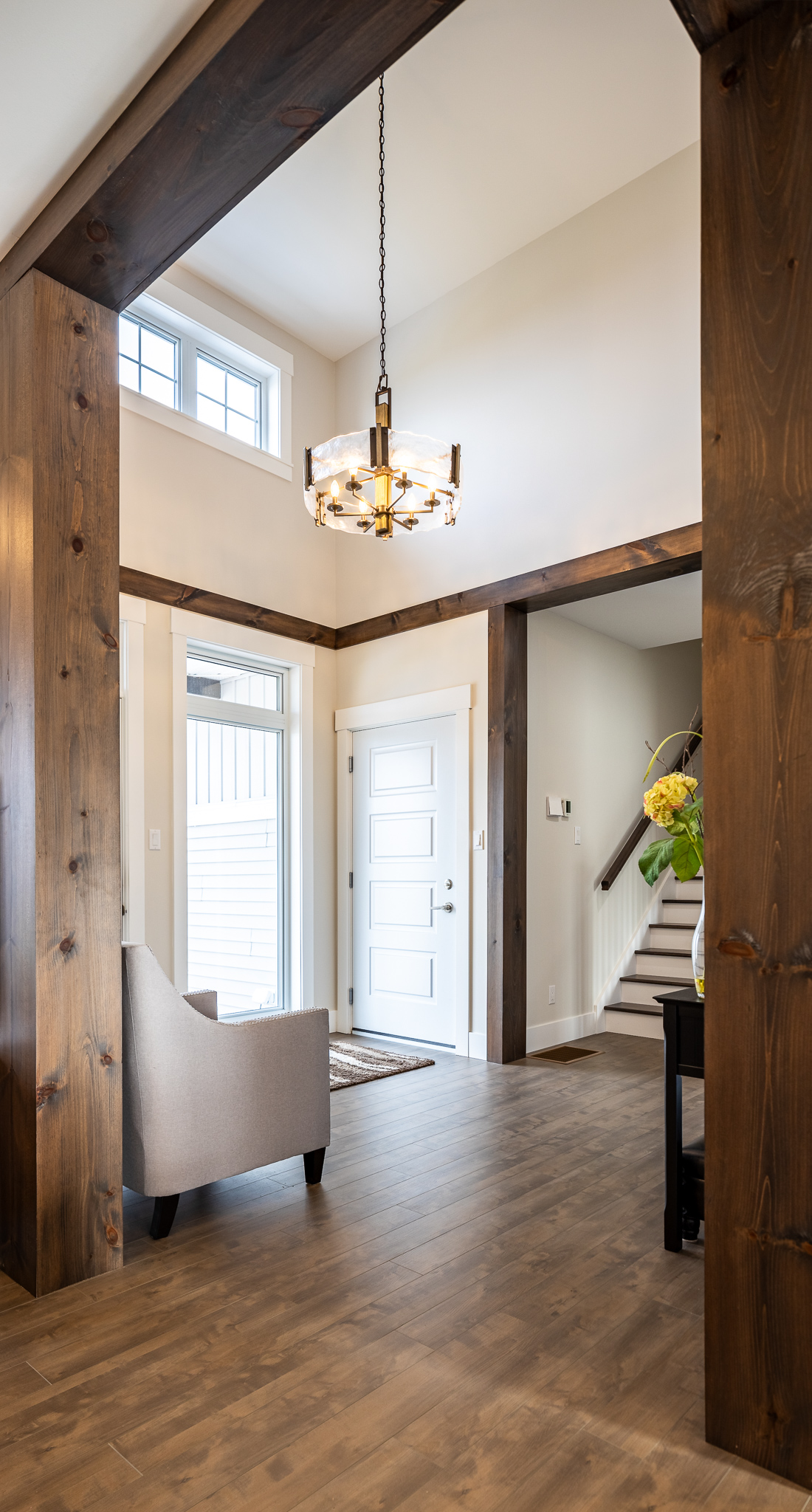 Bright entryway of a home with high ceiling and wooden beams. Features a modern chandelier, white door, upholstered chair, and stairs to the right. Warm and inviting atmosphere.