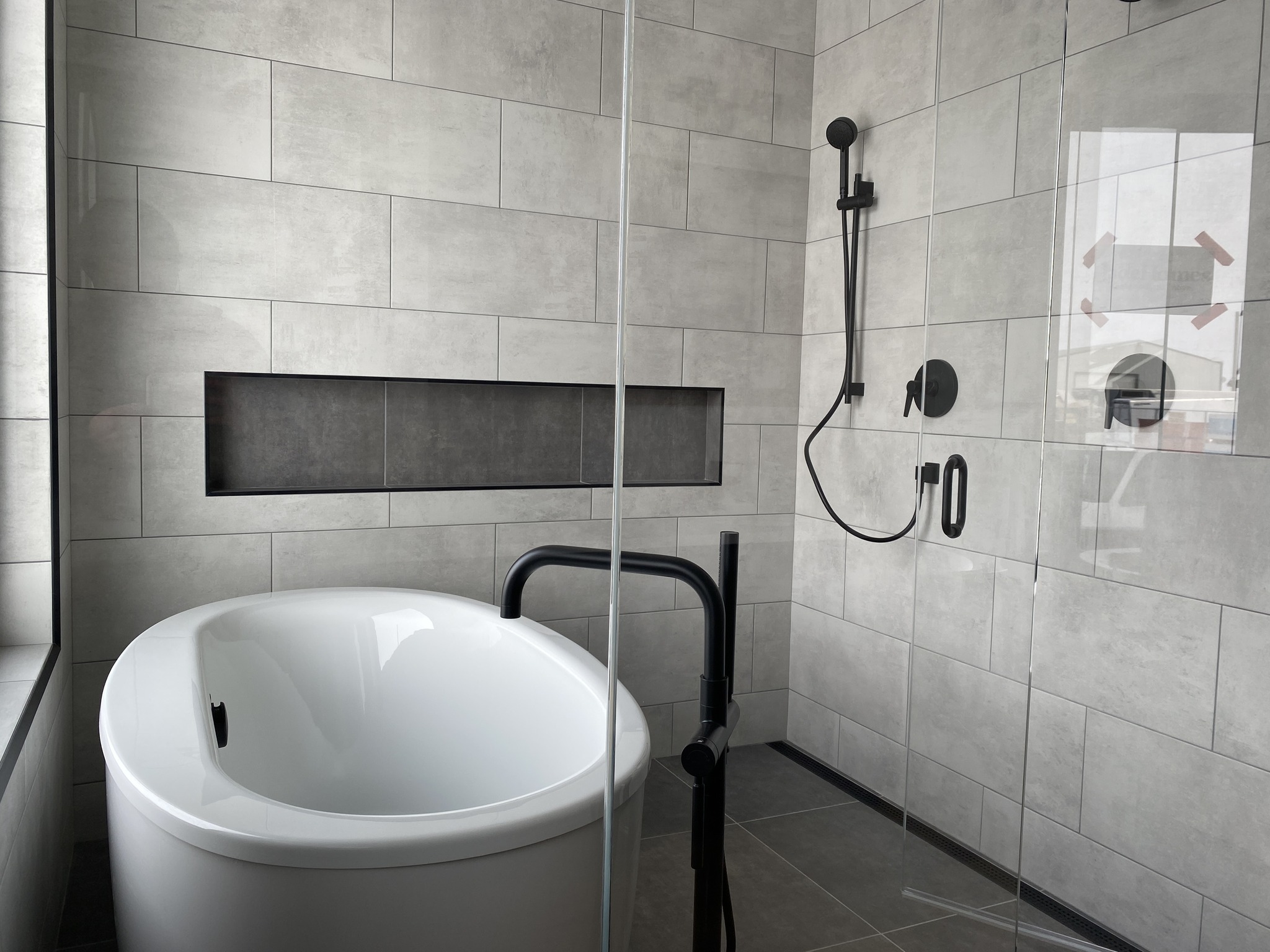 Modern bathroom with gray tiled walls, featuring a white freestanding tub. Black fixtures and a glass shower enclosure add a sleek, minimalist touch.