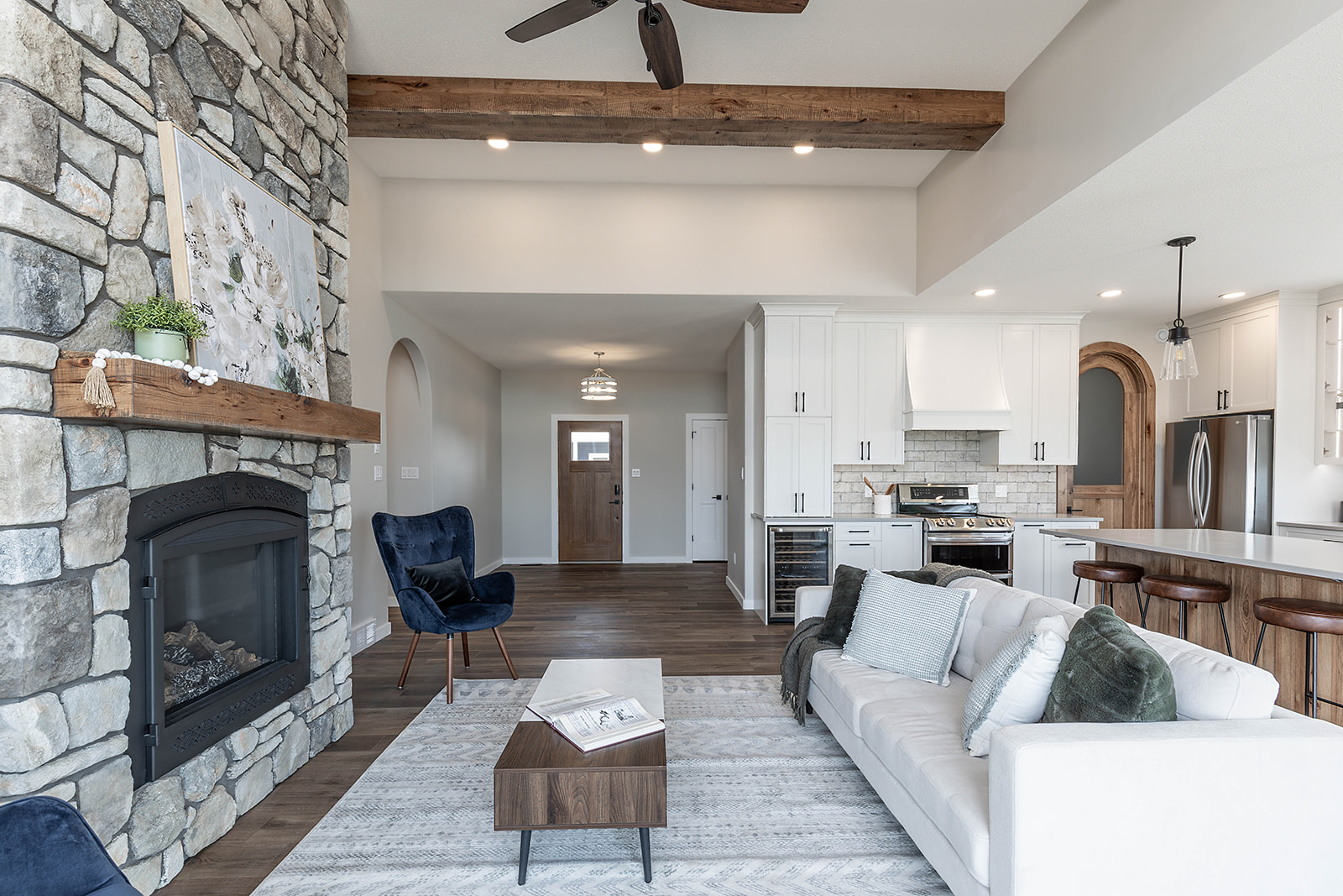 Spacious living room with stone fireplace, blue armchair, white couch, and modern kitchen. Cozy, contemporary atmosphere with wood accents.