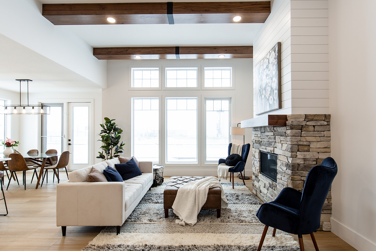 Modern living room with high ceilings, wood beams, large windows, stone fireplace. Features a white sofa, blue chairs, and a cozy dining area.