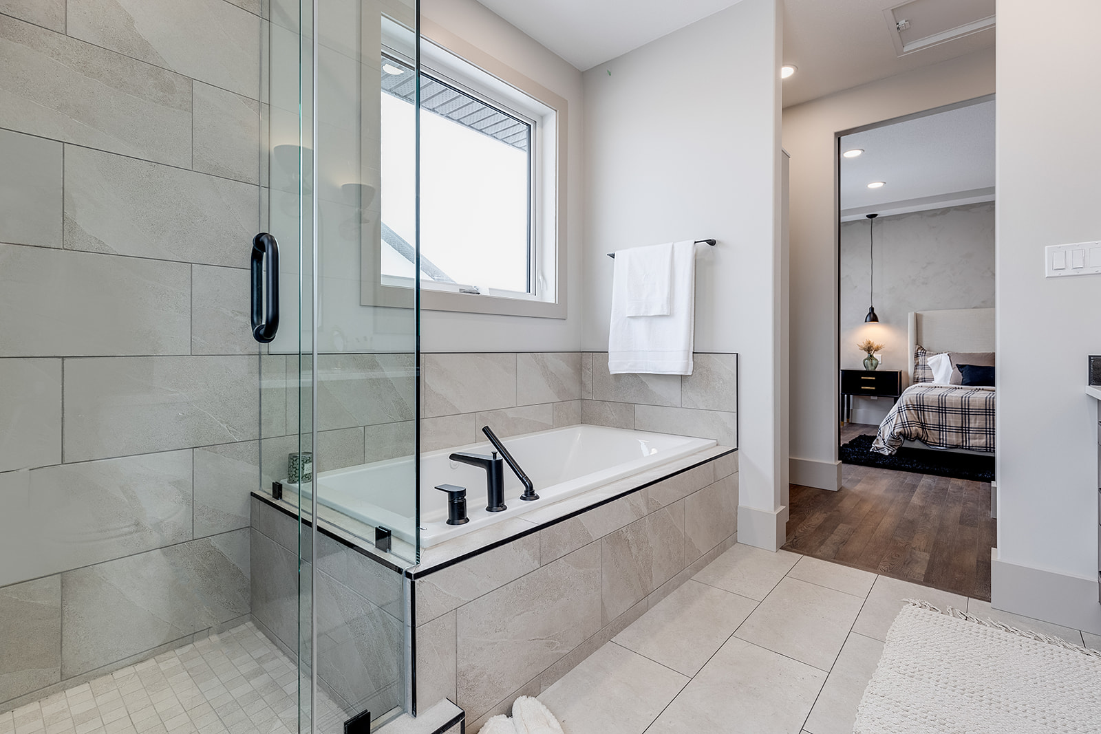 Modern bathroom featuring a glass shower, a rectangular bathtub with black fixtures, and a window. A doorway reveals a cozy bedroom with wood flooring.