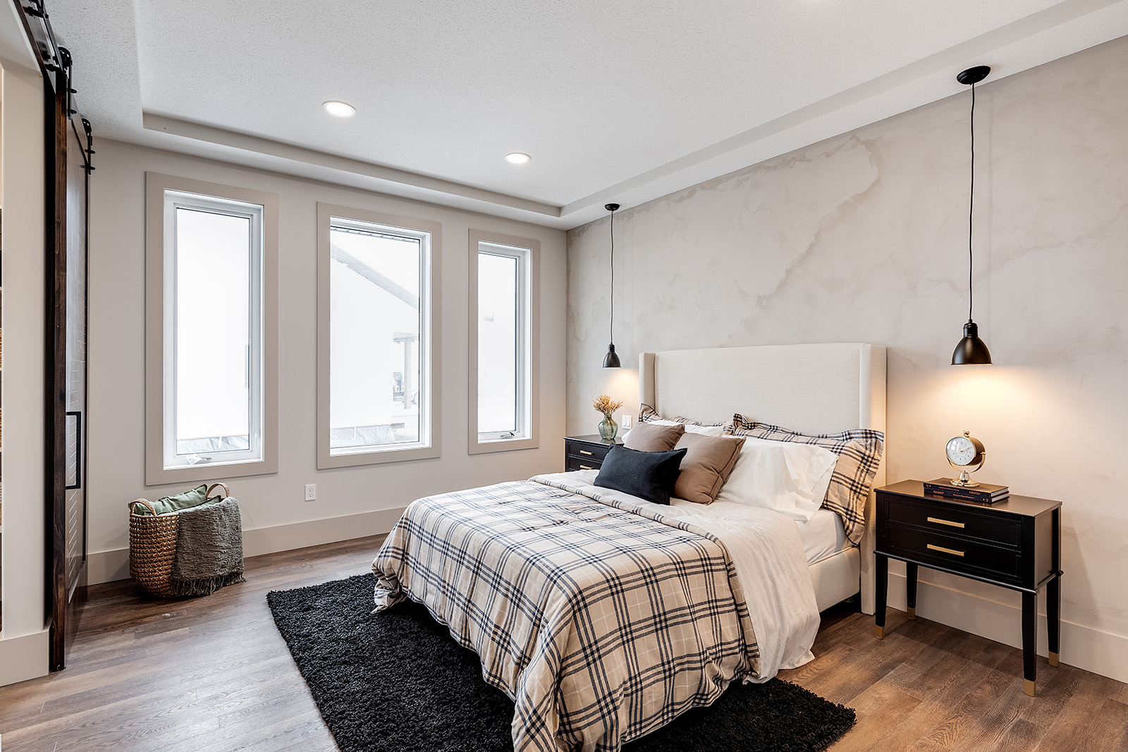 Modern bedroom with a cozy ambiance. Features a plaid bedspread, black rug, bedside tables with lamps, and three tall windows, creating a bright, inviting space.