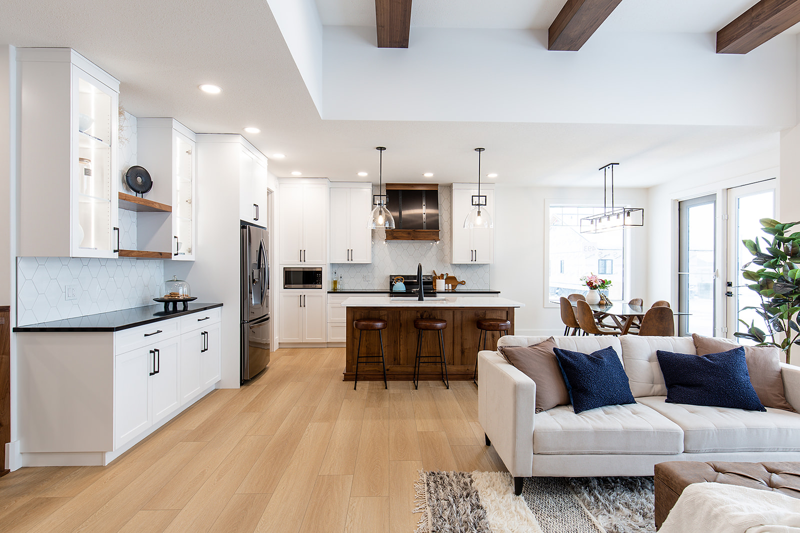 Modern open-plan kitchen and living area with white cabinets, a wooden island, beige sofa, blue cushions, dining table, and pendant lights. Bright and inviting.
