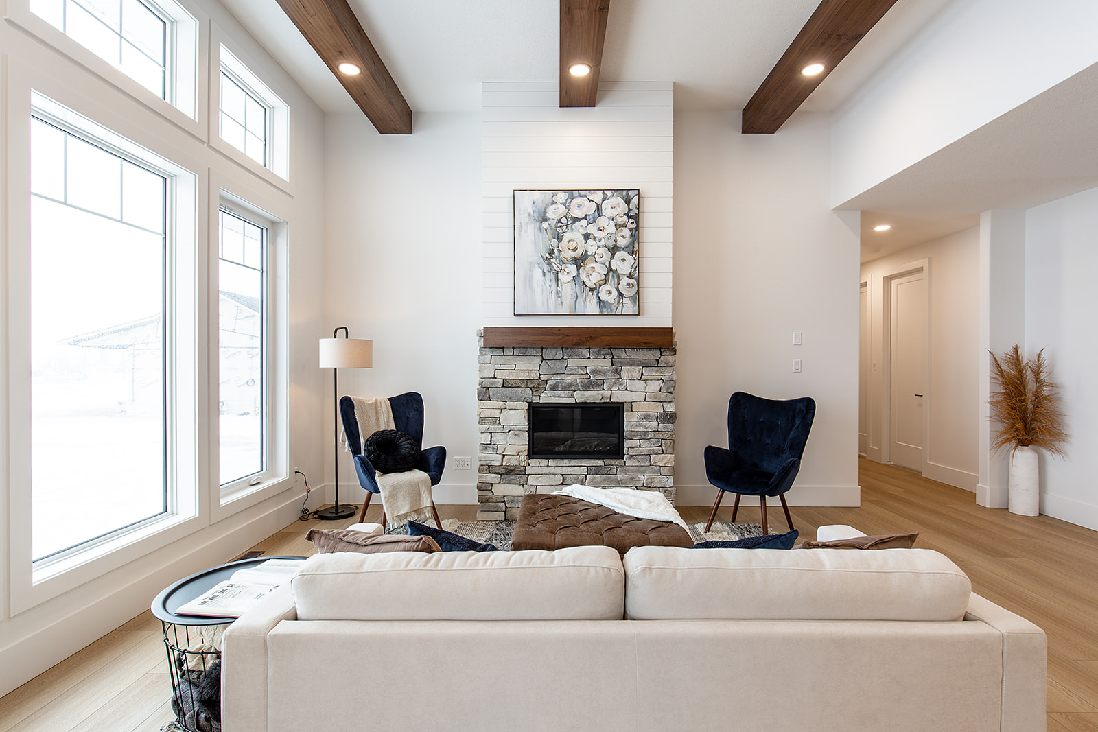 Modern living room with high ceilings, exposed wooden beams, and large windows. Features a stone fireplace, blue velvet chairs, and a beige sofa. Cozy and inviting ambiance.