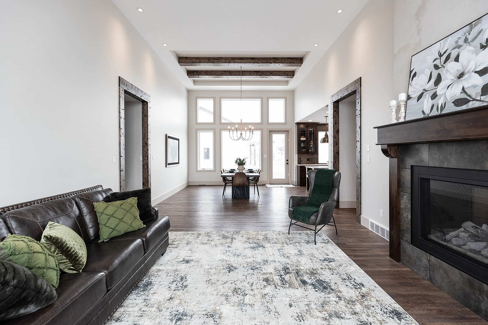 Modern living room with a dark leather sofa, green cushions, and a textured rug. Features a fireplace, dining area, chandelier, and large windows, creating an airy, inviting atmosphere.