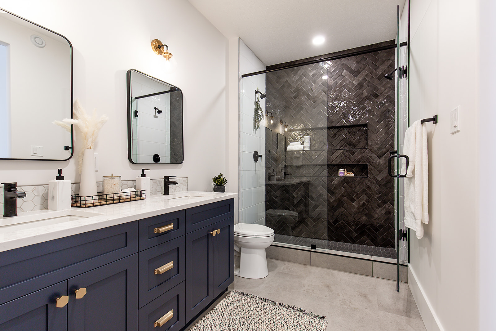 Modern bathroom with navy double vanity, dual mirrors, and gold fixtures. Glass shower has herringbone tile; soft lighting creates a relaxed ambiance.