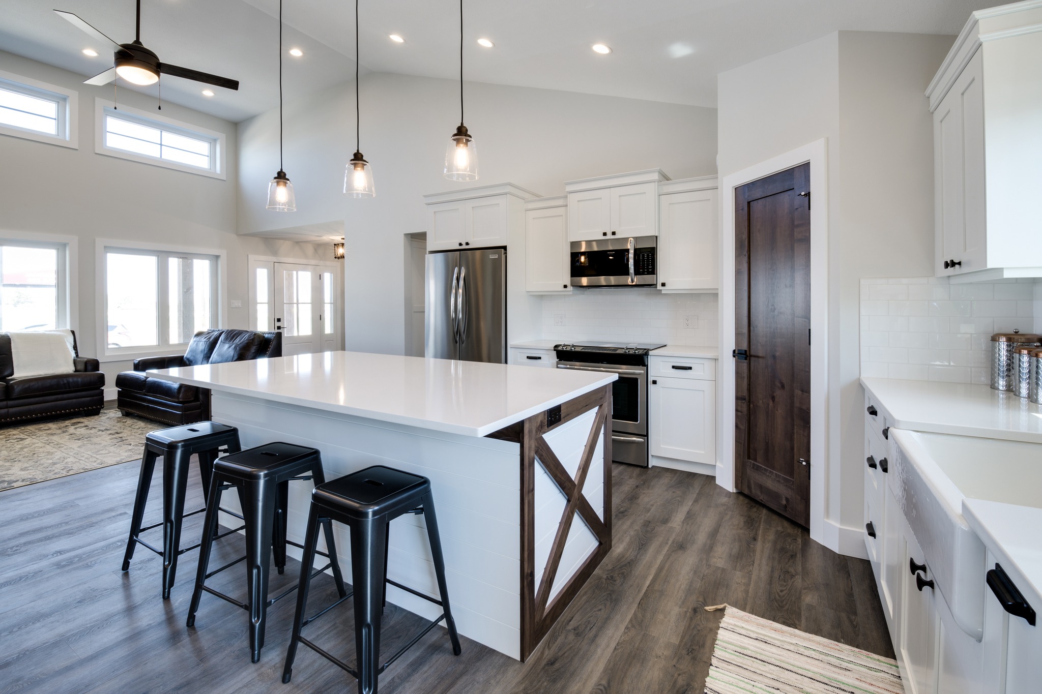 Modern kitchen with white cabinetry, dark wood accents, and stainless steel appliances. Features a large island with black stools and pendant lights. Bright and airy space with high ceilings and wood floors.