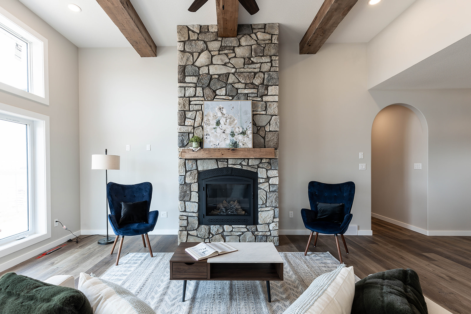 Cozy living room with a stone fireplace, flanked by two blue velvet chairs. Warm light filters through large windows, enhancing the rustic ambiance.