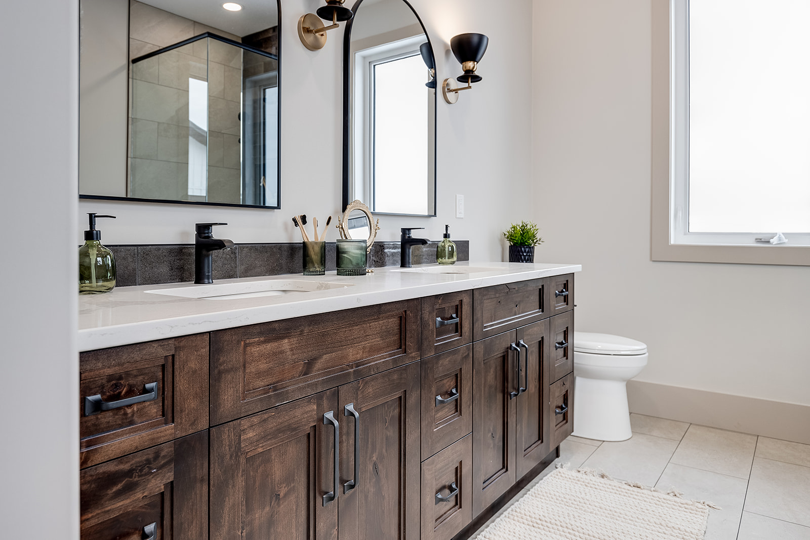 Modern bathroom with a wooden double vanity, black fixtures, and a large mirror. It has a bright, clean aesthetic with plants and a wall sconce.