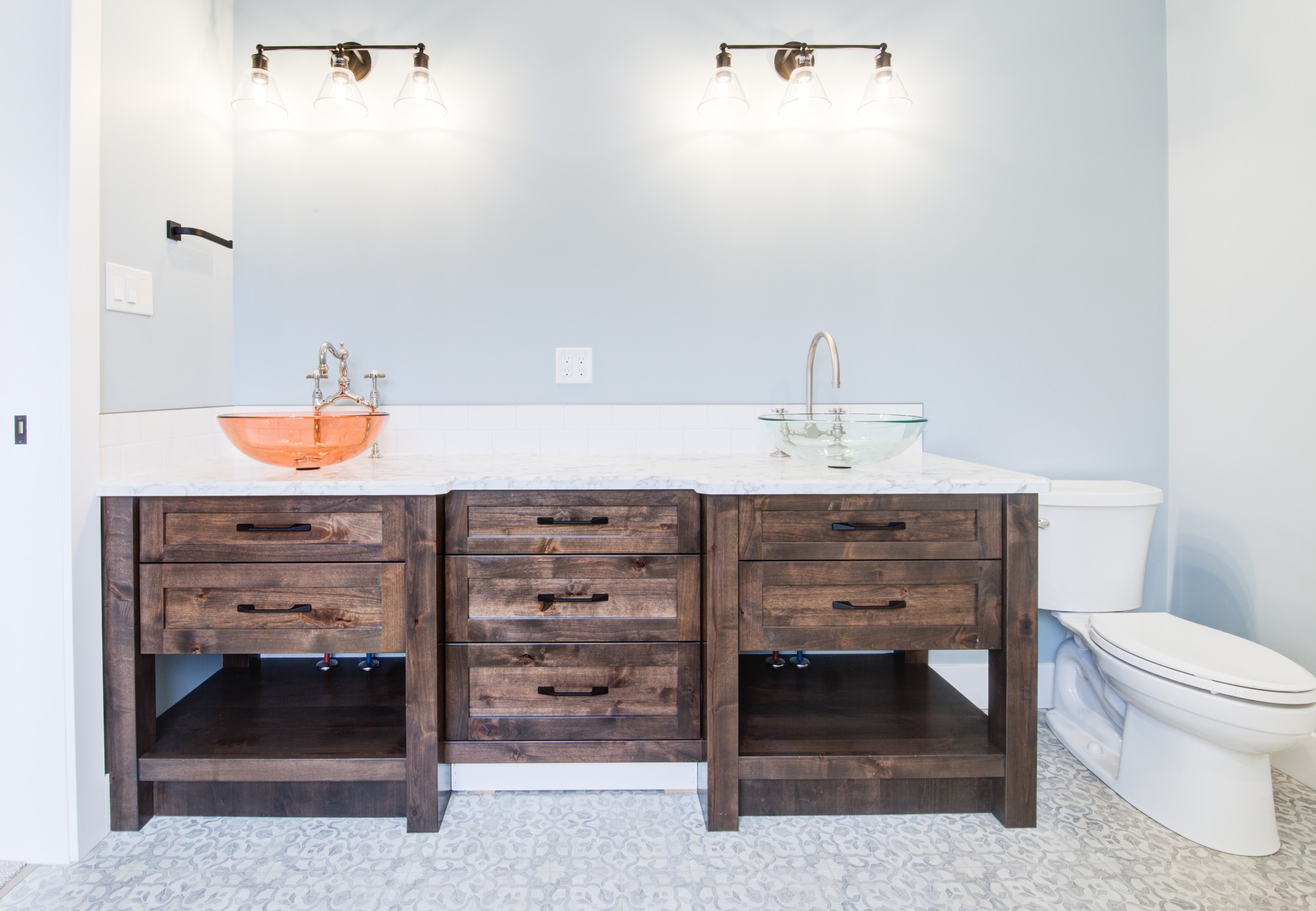 Spacious bathroom with a wooden double vanity. Copper and glass vessel sinks sit beneath modern lights. White toilet on the right. Calm, minimalist ambiance.