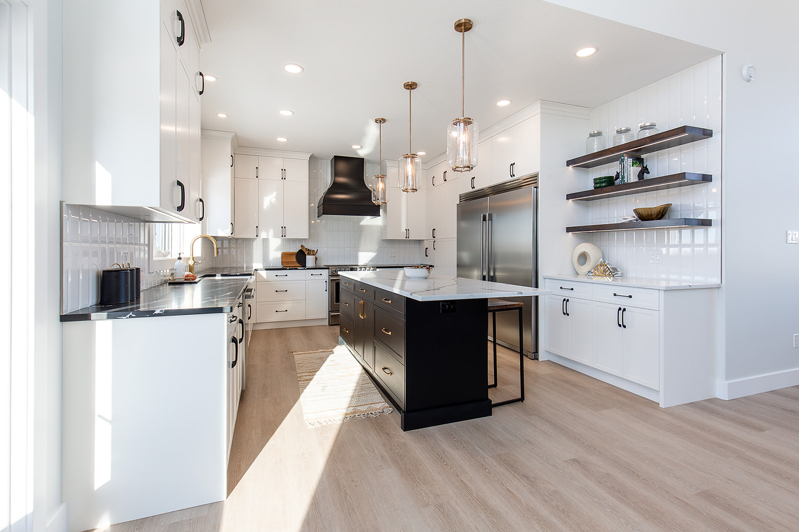 Modern kitchen with white cabinets, wood floor, and a central black island. Pendant lights hang above, and open shelves display decor. Bright and inviting.