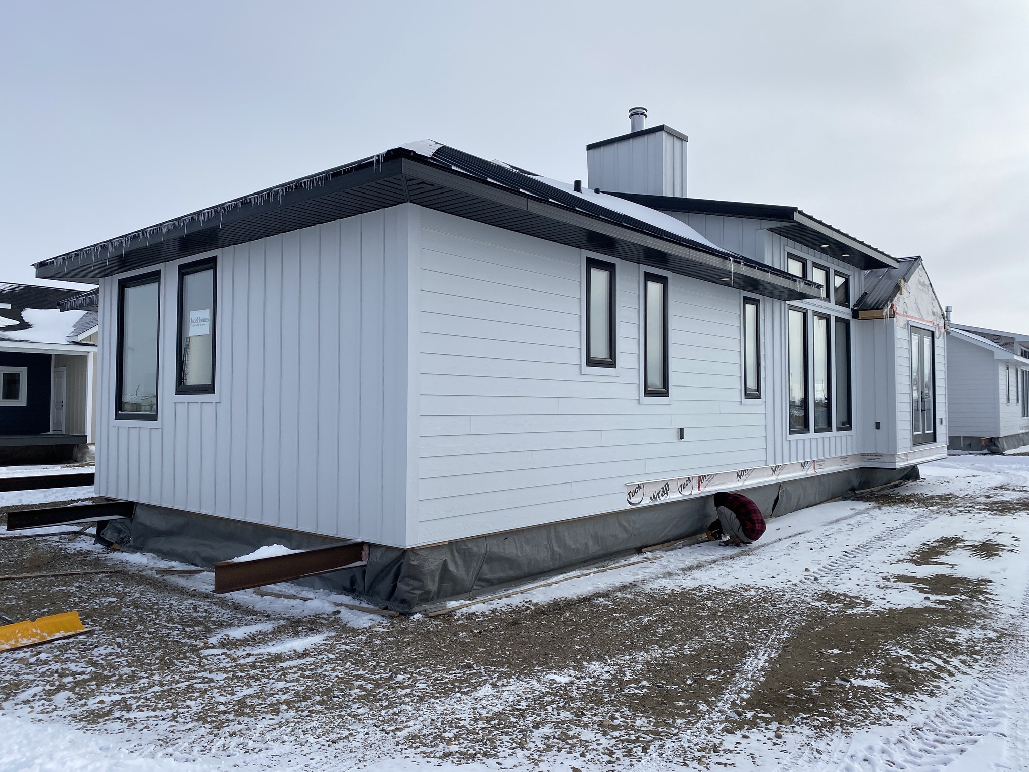 A modern, white, modular home sits on a snowy lot under a gray sky. The house features large windows and a chimney, conveying a new, unfinished feel.
