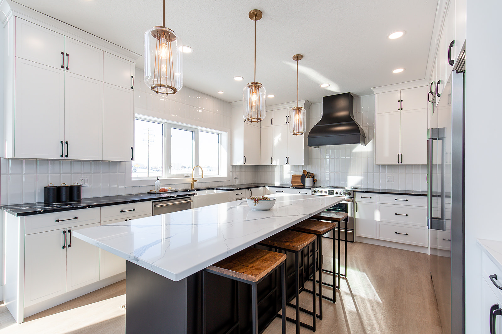 Spacious modern kitchen with white cabinets, large island with wooden stools, pendant lights, black range hood, and bright, natural lighting.