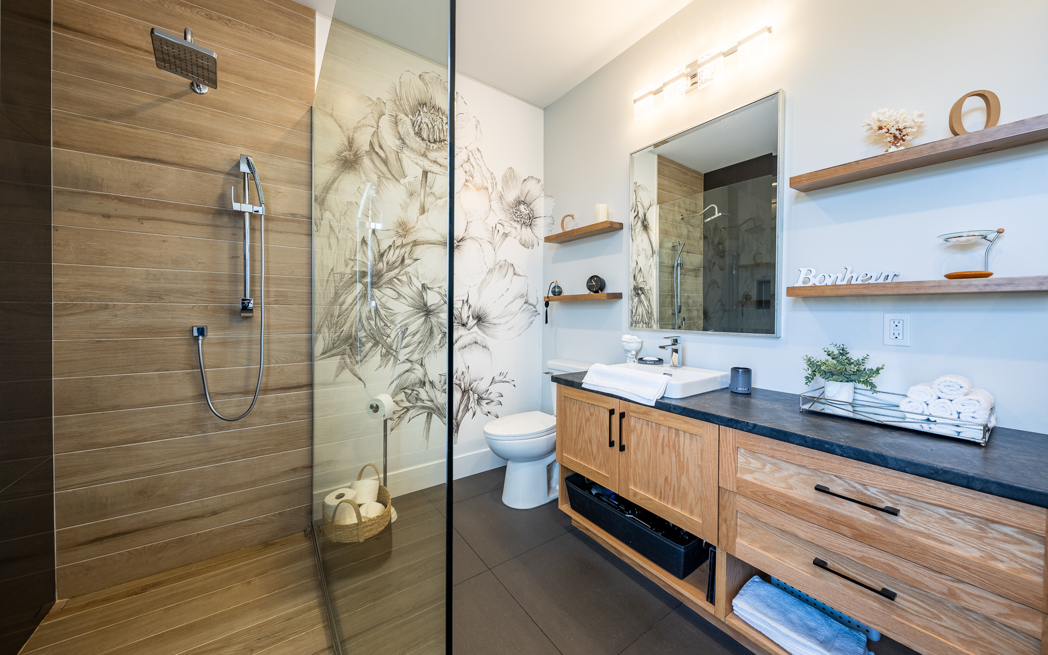 Modern bathroom with a wooden theme. Glass shower on the left, floral mural, and vanity with drawers on the right. Shelves hold decor and essentials. Warm, serene ambiance.