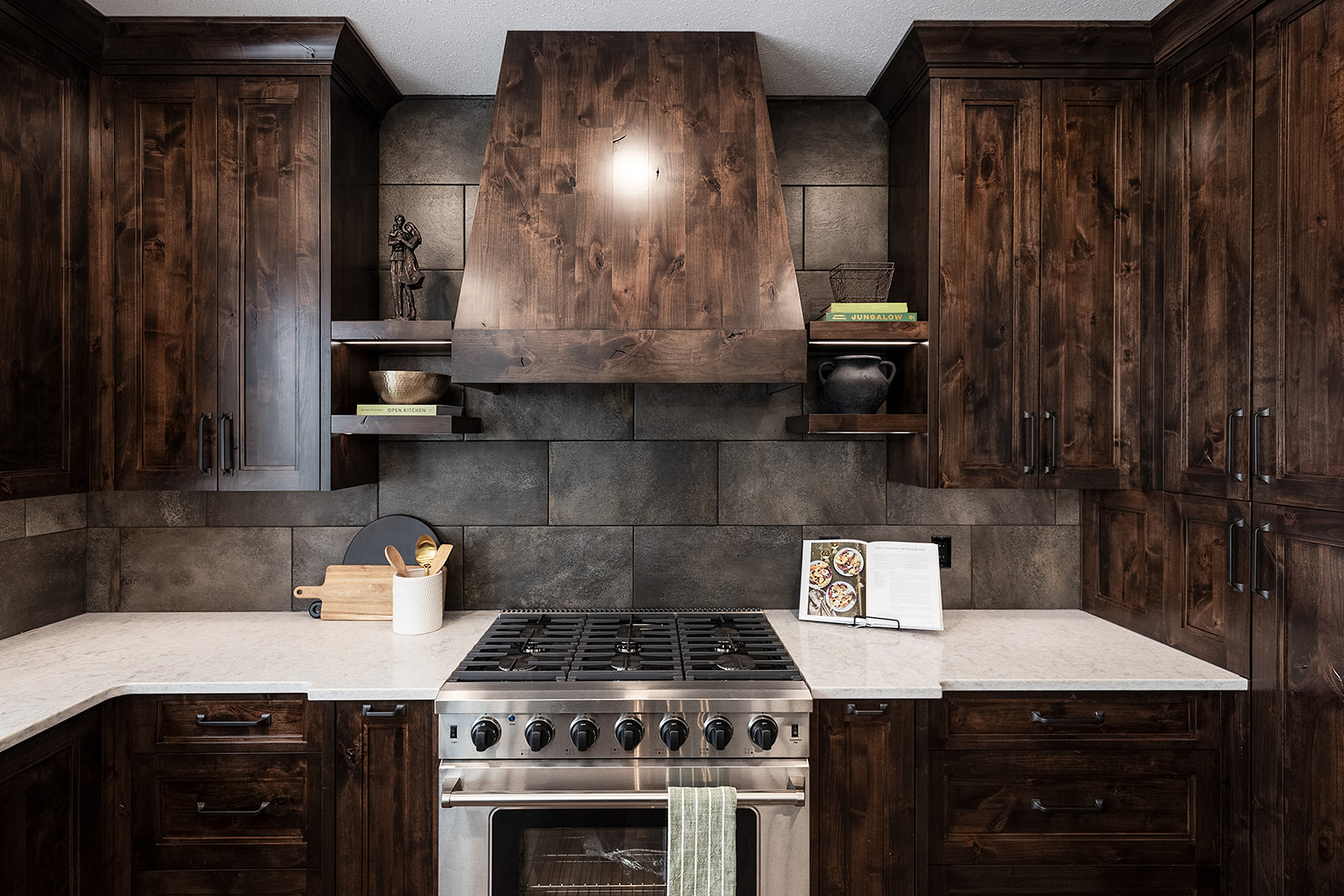 Rustic kitchen with dark wooden cabinets, stone backsplash, and a stainless-steel stove. Open shelves hold books and dishes, creating a cozy ambiance.