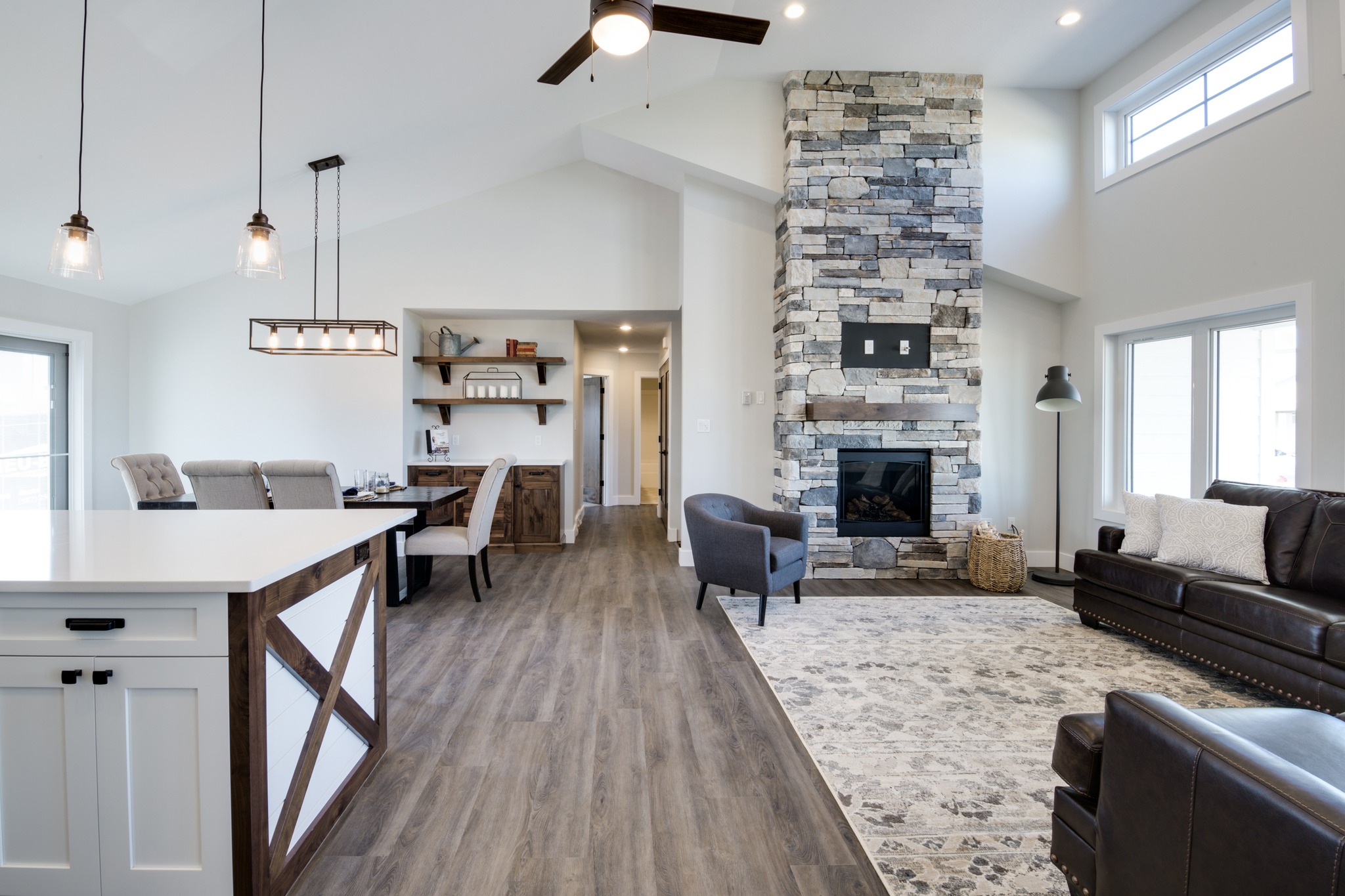 Spacious open-concept living room with high ceilings, stone fireplace, and modern kitchen. Hardwood floors and cozy, neutral furnishings create a warm ambiance.