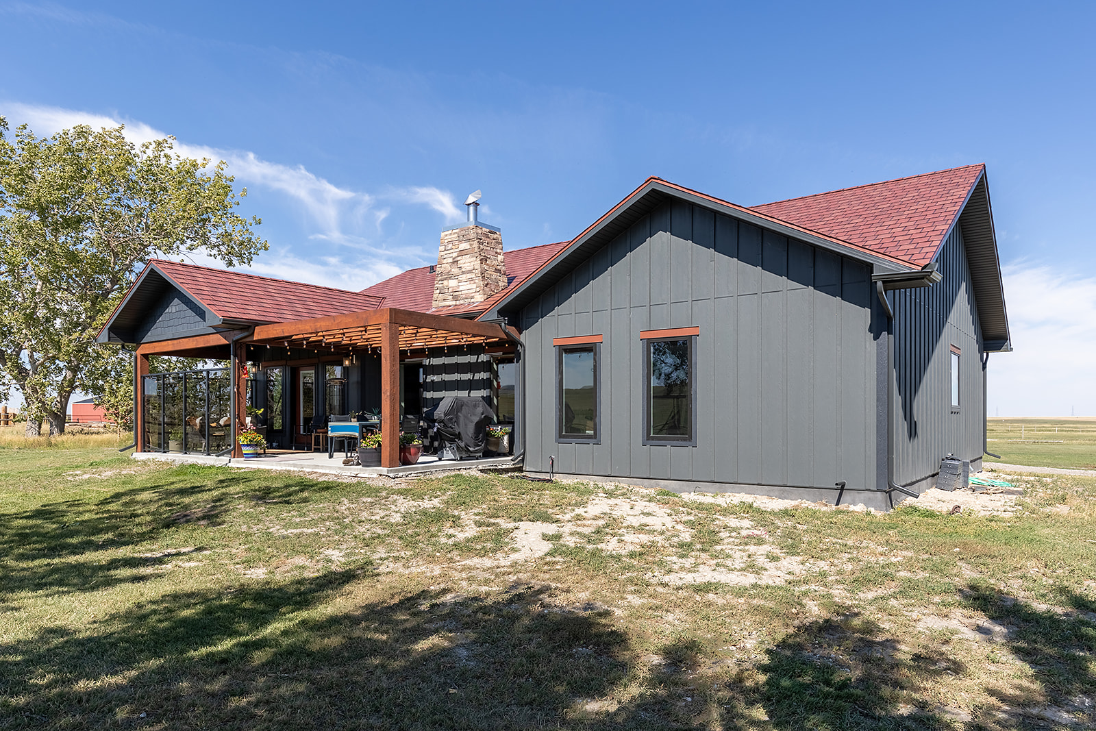 Modern house with dark gray siding and red roof, featuring a stone chimney and covered patio. Surrounded by grass and trees under a clear blue sky.