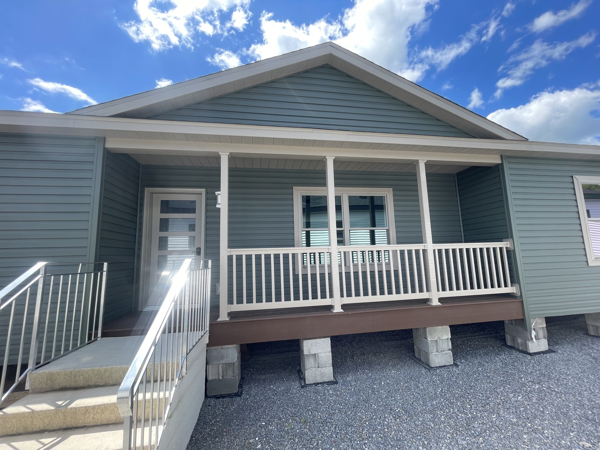 Single-story house with green siding, front porch, and white railing. A short ramp with metal handrails leads to the door. Clear blue sky above.