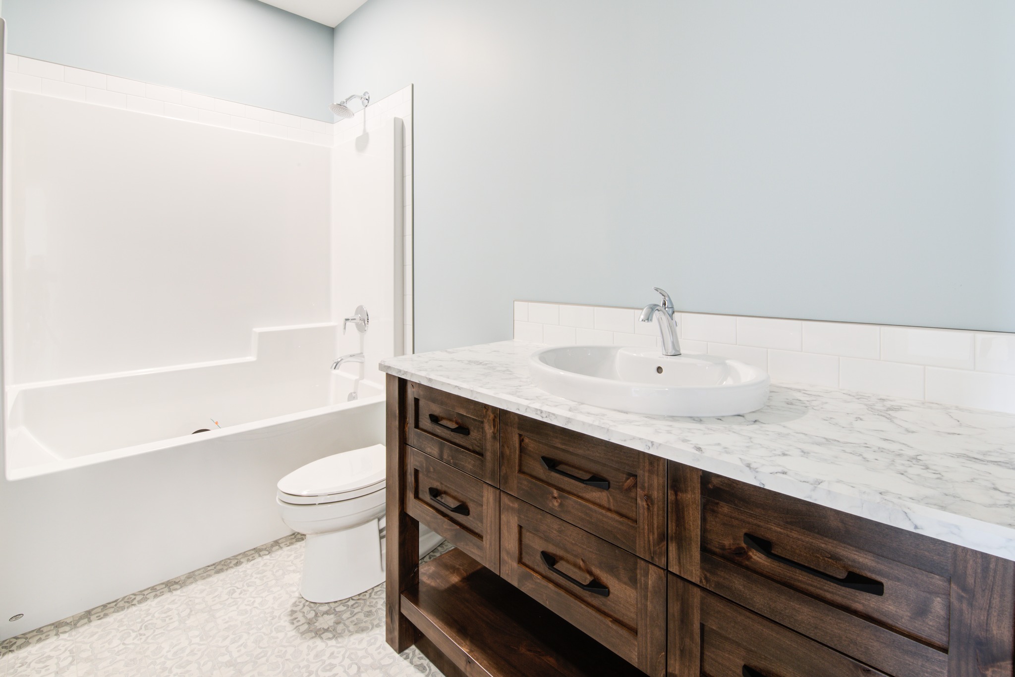 A clean, modern bathroom with light blue walls features a white bathtub, a toilet, and a wooden vanity with a marble countertop, exuding a calm and minimalist vibe.