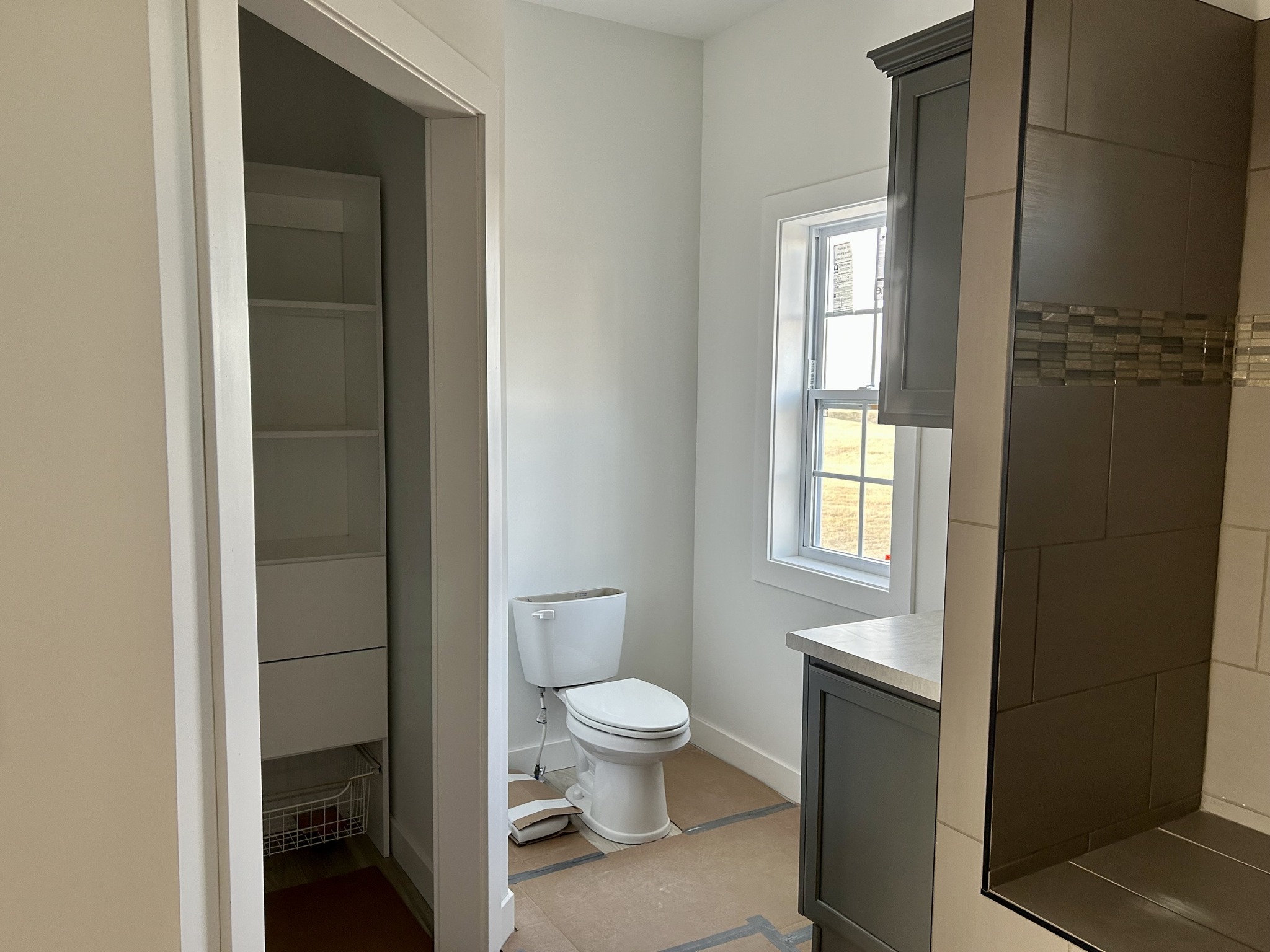 A modern bathroom with a white toilet, light grey walls, a narrow window, and adjacent dark-colored cabinetry and tiled shower. The atmosphere is clean and minimalistic.