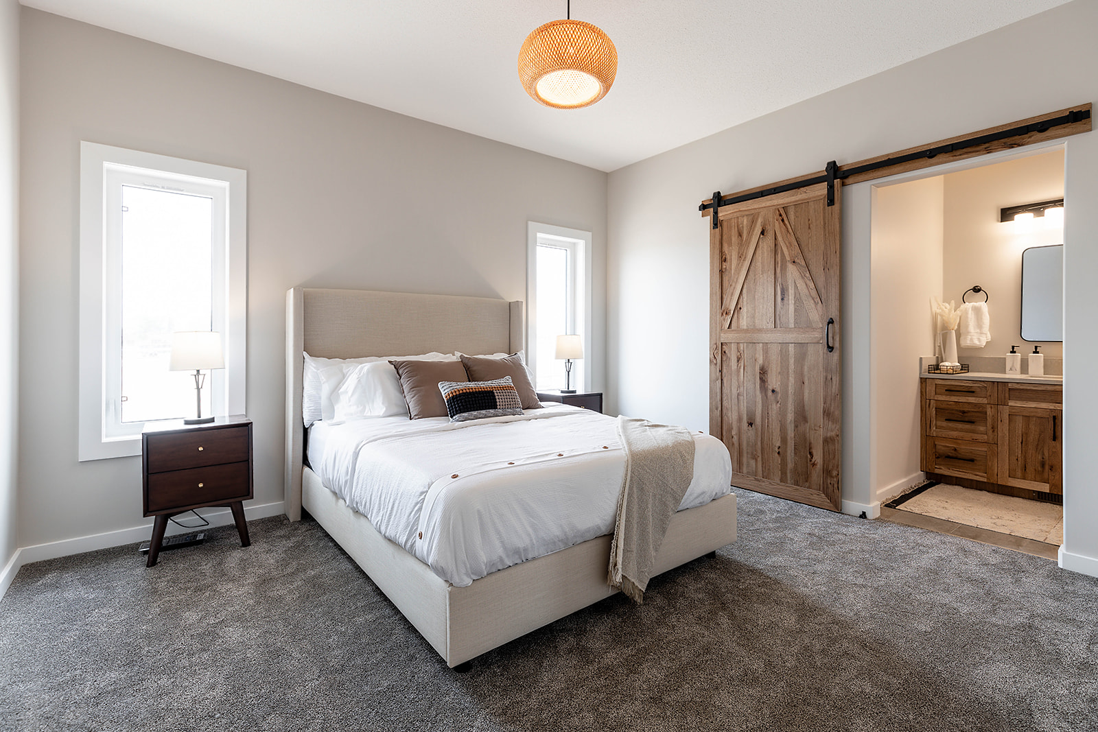 Modern bedroom with a cozy vibe, featuring a plush bed, soft gray carpet, wooden nightstands, and a sliding barn door leading to a warm-lit bathroom.