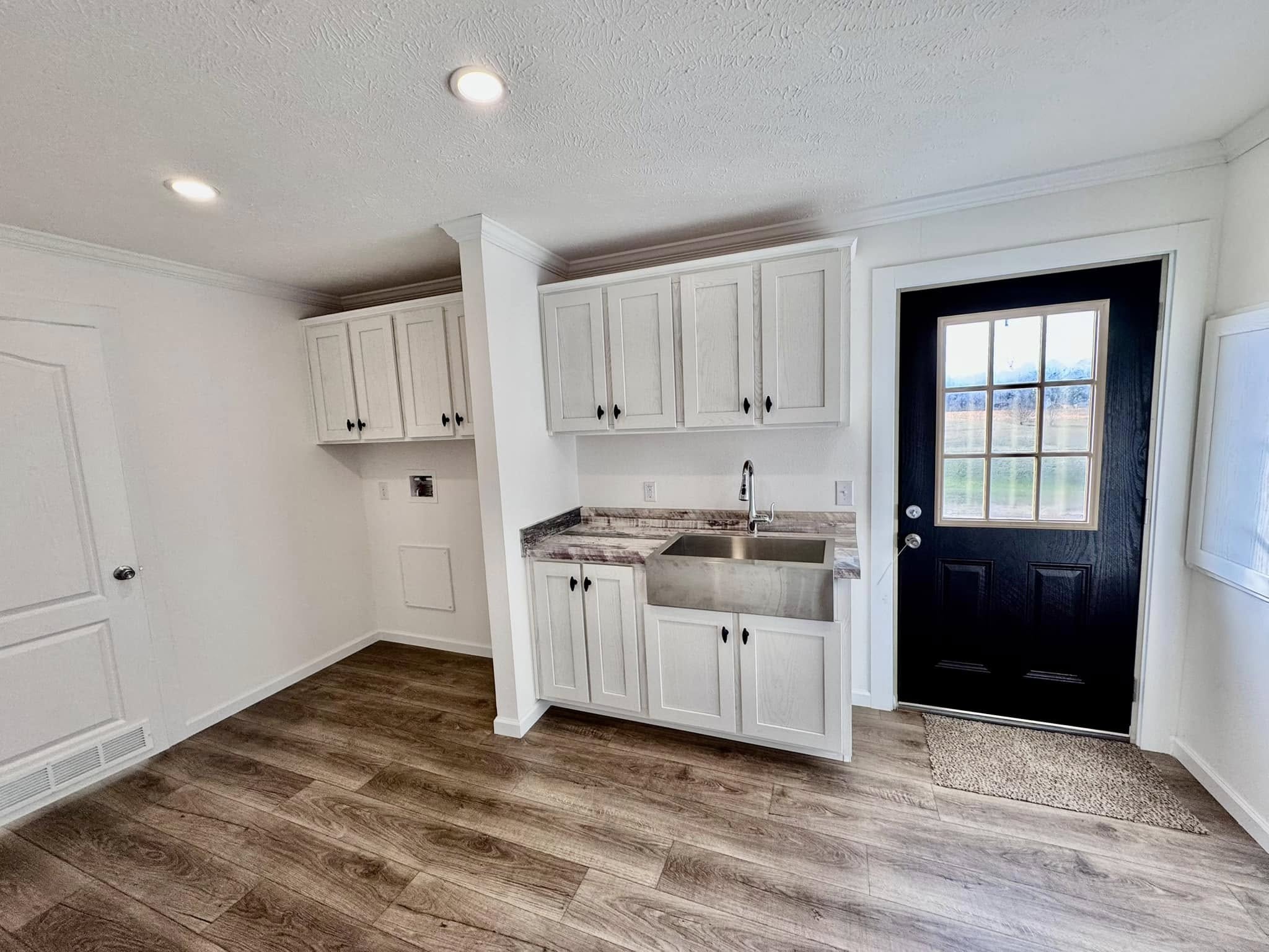 Modern utility room with white cabinetry, stainless steel sink, and dark wood floor. A black door with glass panels opens to the outside. Bright and tidy.