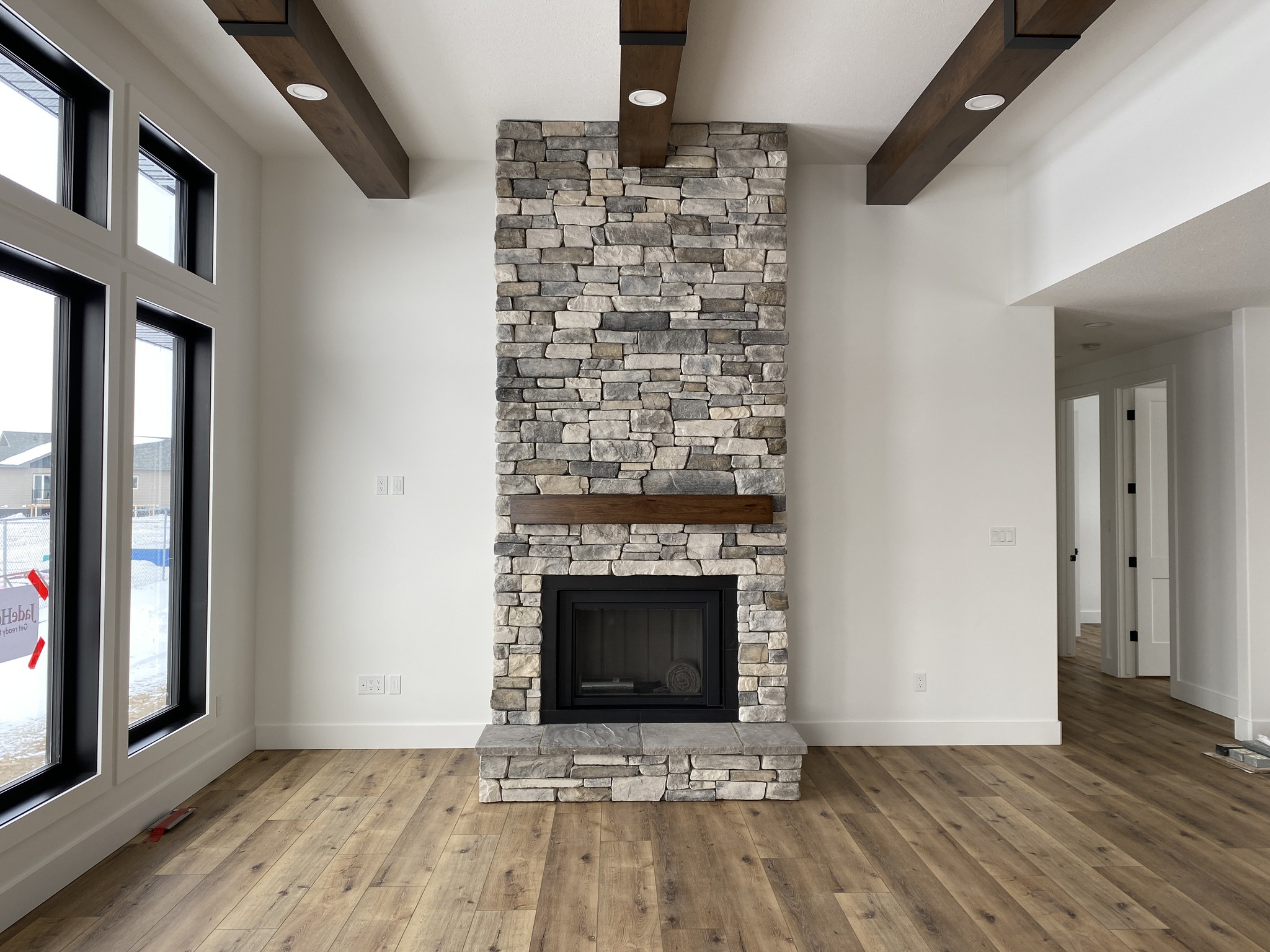 Modern living room with a stacked stone fireplace as the focal point, featuring wooden beams and light wood flooring. Large windows provide natural light.