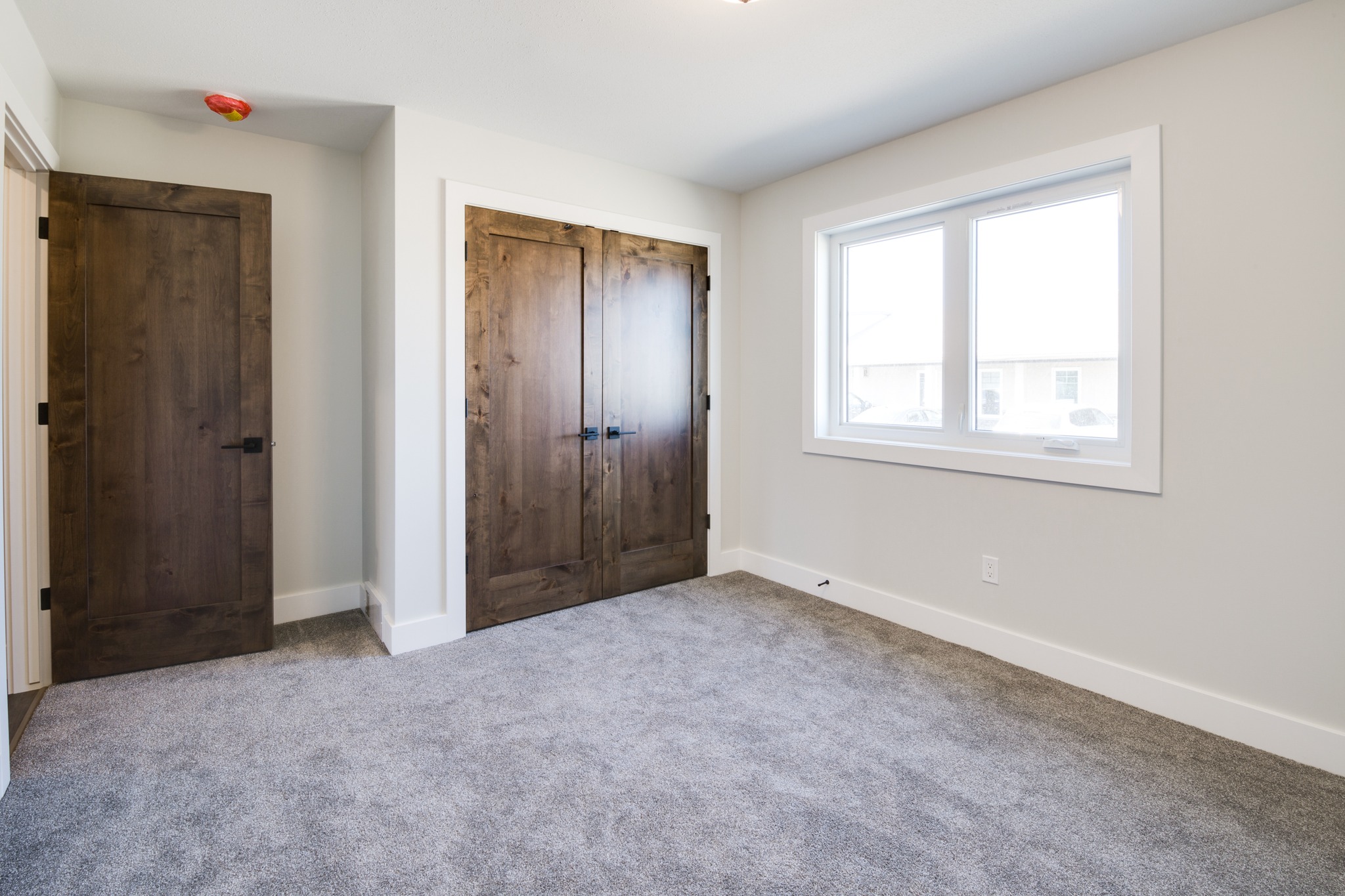 A small, empty room with beige walls and gray carpet. It features a large window with white trim and dark wood closet doors, evoking a calm feel.