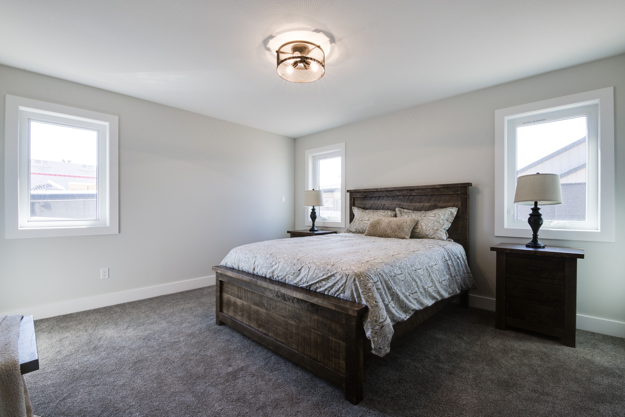 Bedroom with a cozy ambiance, featuring a wooden bed and matching nightstands, two windows, gray carpet, and a round ceiling light fixture.