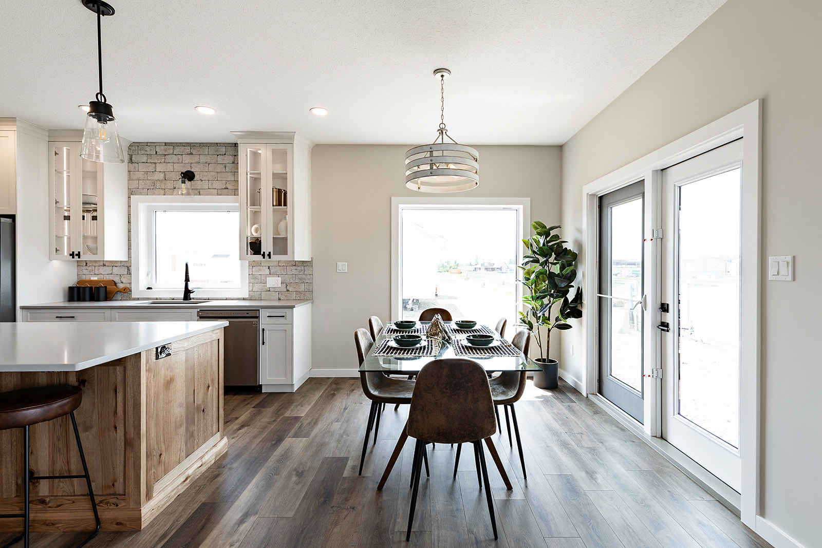 Modern kitchen and dining area with wooden floors, a light wooden island, and white cabinetry. A set dining table, large windows, and a potted plant add brightness.