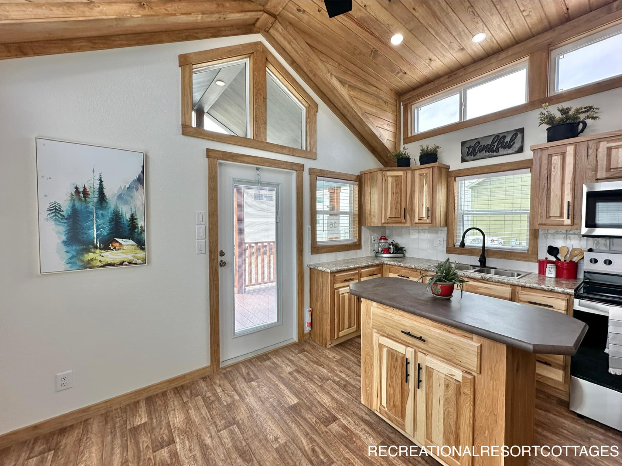 Cozy kitchen with wooden ceiling and floors, featuring natural wood cabinets, a central island, wall art, and windows allowing ample natural light.