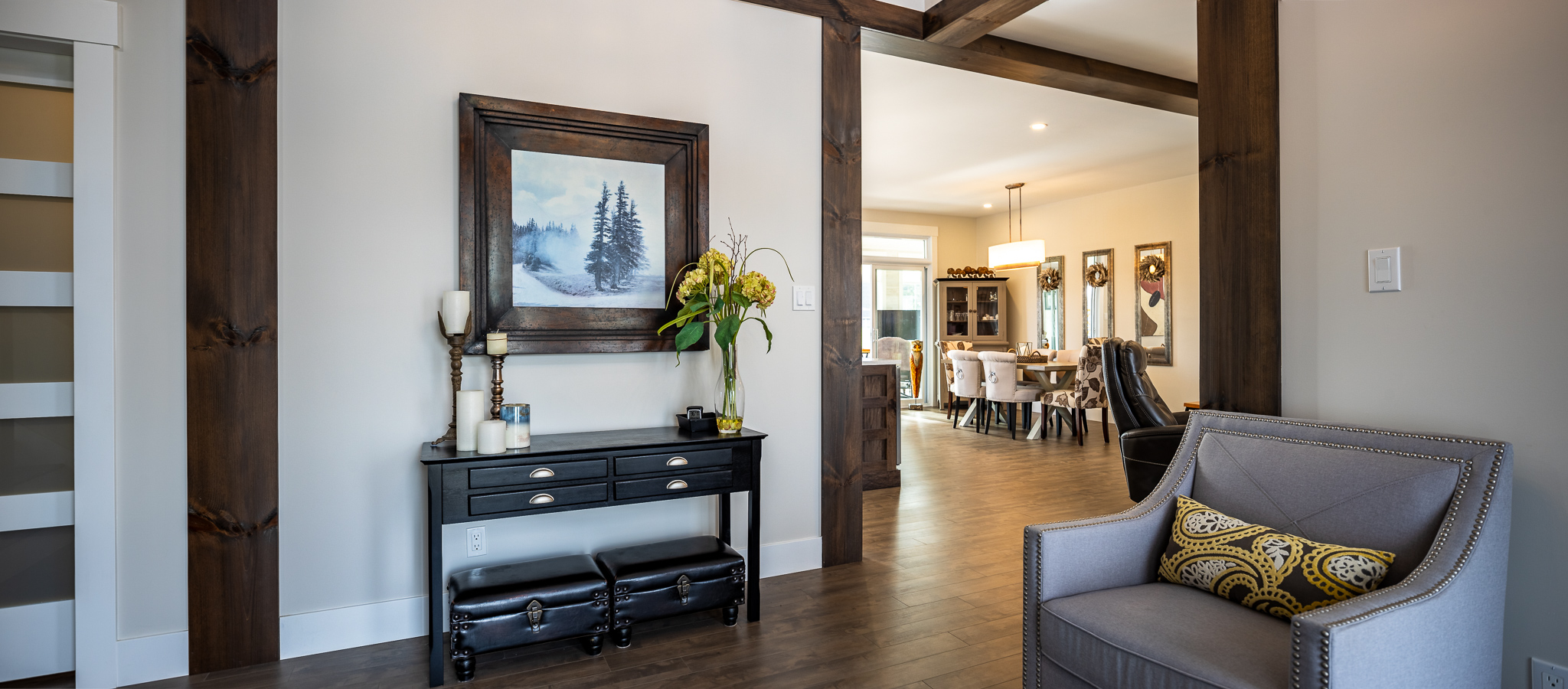 Cozy living room with a plush gray armchair, patterned cushion, and a black console table. A framed landscape art piece and potted plant above evoke tranquility.